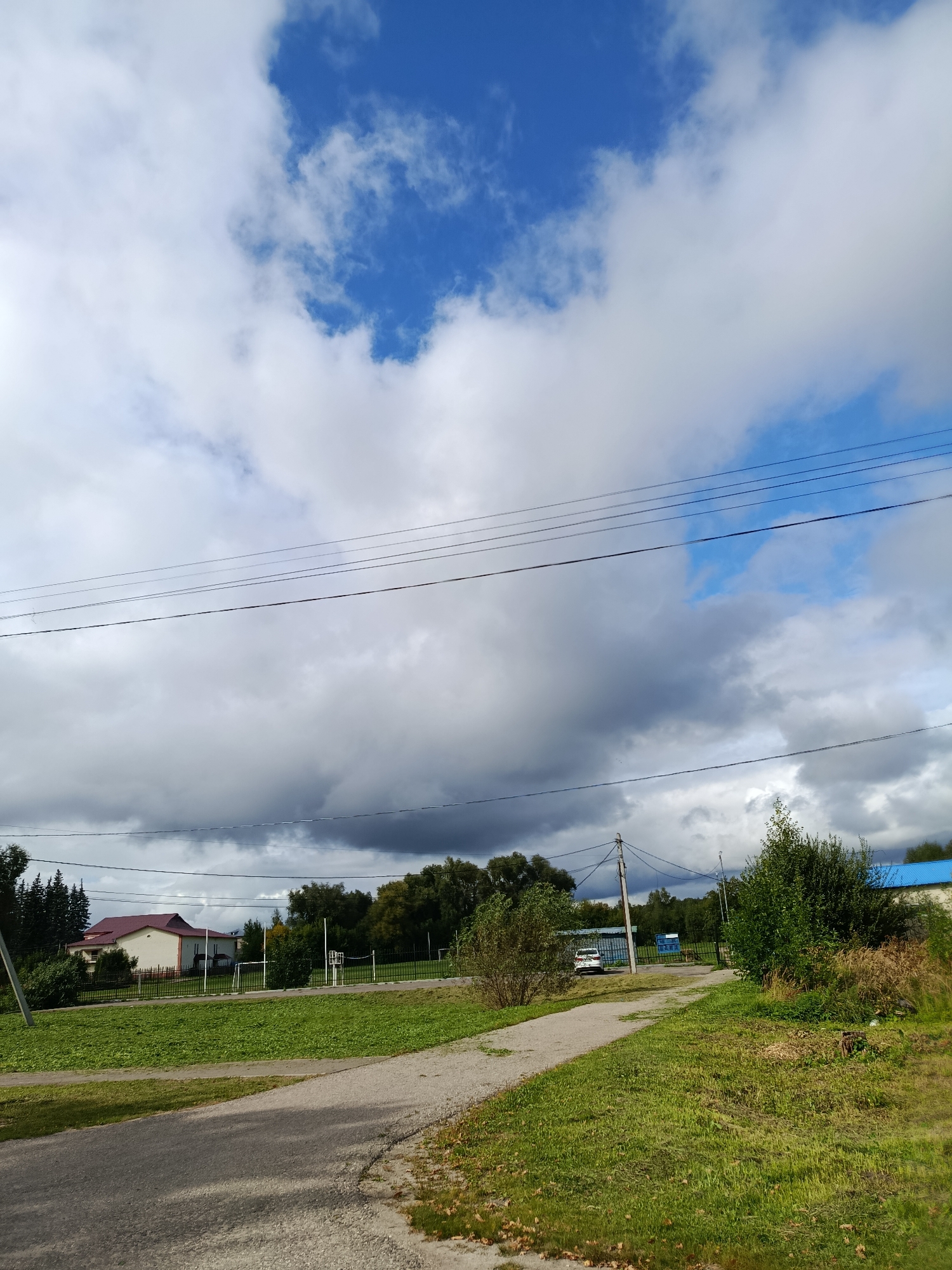 The image depicts an outdoor scene with a cloudy sky. In the foreground, a grey asphalt path leads into the frame. To the right of the path is a grassy area with some dry vegetation. Further into the scene, a wider paved area or road is visible, with a white car parked on the left side. On the right, there is a building with a blue roof and a large green bush. A metal fence runs across the middle ground, separating the paved area from a lawn and a building with a red roof on the left. Beyond the fence, there are trees and a sports field with a goal. Utility poles and power lines are present throughout the scene, crossing the sky. The location is identified as Verea, Russia.