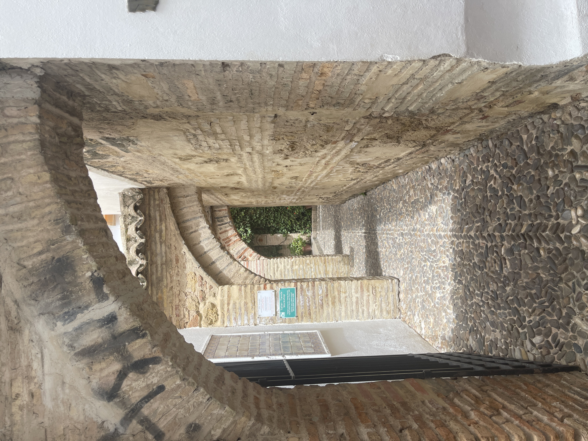 The image depicts an upward view through a narrow, arched passageway in what appears to be an old European city. The passageway is constructed from a mix of materials, including rough-hewn stone, brickwork, and possibly concrete. The walls on the right are clad in small, rounded pebbles, creating a textured surface. The ceiling is a rough, aged brick arch. Sunlight casts shadows, suggesting it is daytime, and the overall atmosphere is one of historical charm and perhaps a bit of disrepair.
In the distance, an opening reveals a glimpse of lush green foliage, hinting at a courtyard or garden. A sign with Spanish text is visible on the left wall, appearing to be informational or directional. Below the sign, a tiled grate or window is set into the floor or a lower wall. The foreground shows more brickwork and the curve of an arch, with some dark, graffiti-like marks on the left side.
The setting strongly suggests a historic district within Córdoba, Spain, possibly a narrow alleyway or a covered passage leading to an interior space. There are no people or animals visible in the frame. The focus is on the architecture and textures of the passageway. The weather appears to be clear and sunny.