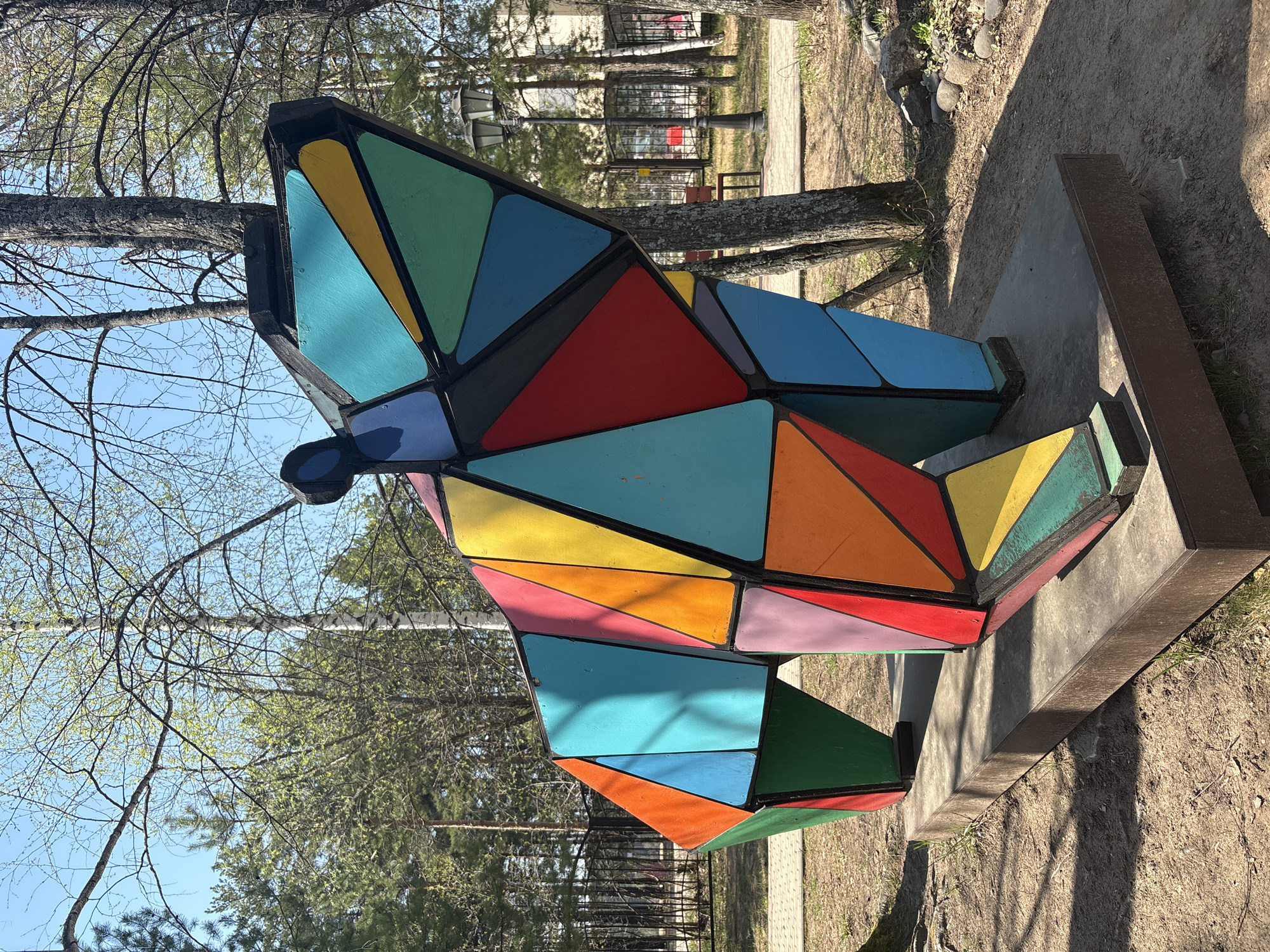 The image shows a large, colorful abstract sculpture located outdoors in Chaikovsky, Russia. The sculpture is composed of geometric, triangular shapes, painted in a variety of bright colors including red, blue, green, yellow, and orange. The shapes are constructed with black borders. The sculpture rests on a concrete base. The background includes trees with bare branches, suggesting early spring or late autumn. In the distance, glimpses of other buildings and structures within a park or public area are visible. The ground is covered with dirt and some grass. The sky is blue, with some sparse clouds.