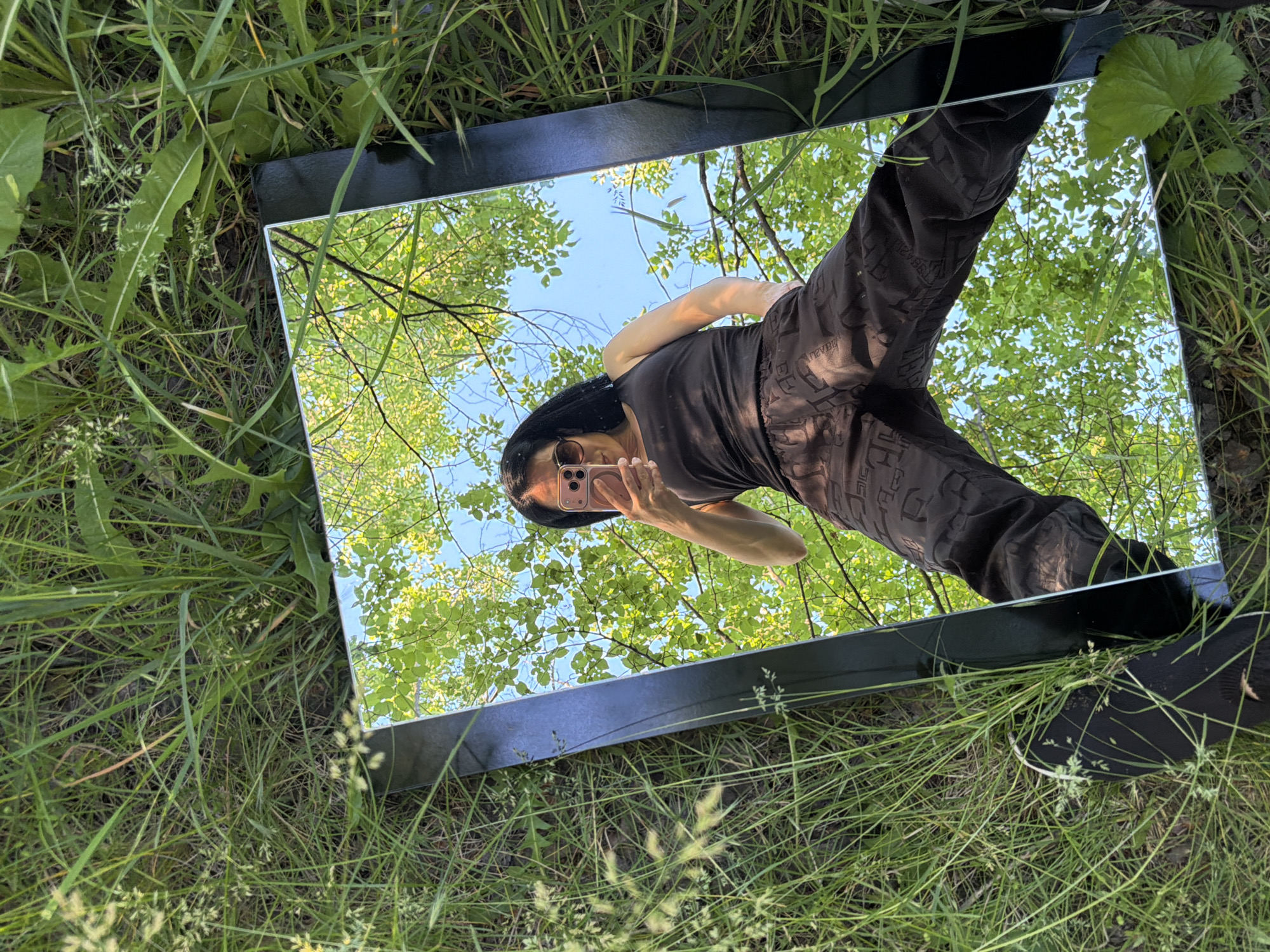 A mirror with a black frame rests on grass. The mirror reflects a woman holding a cell phone, the sky, and trees. The location is not discernible from the image.