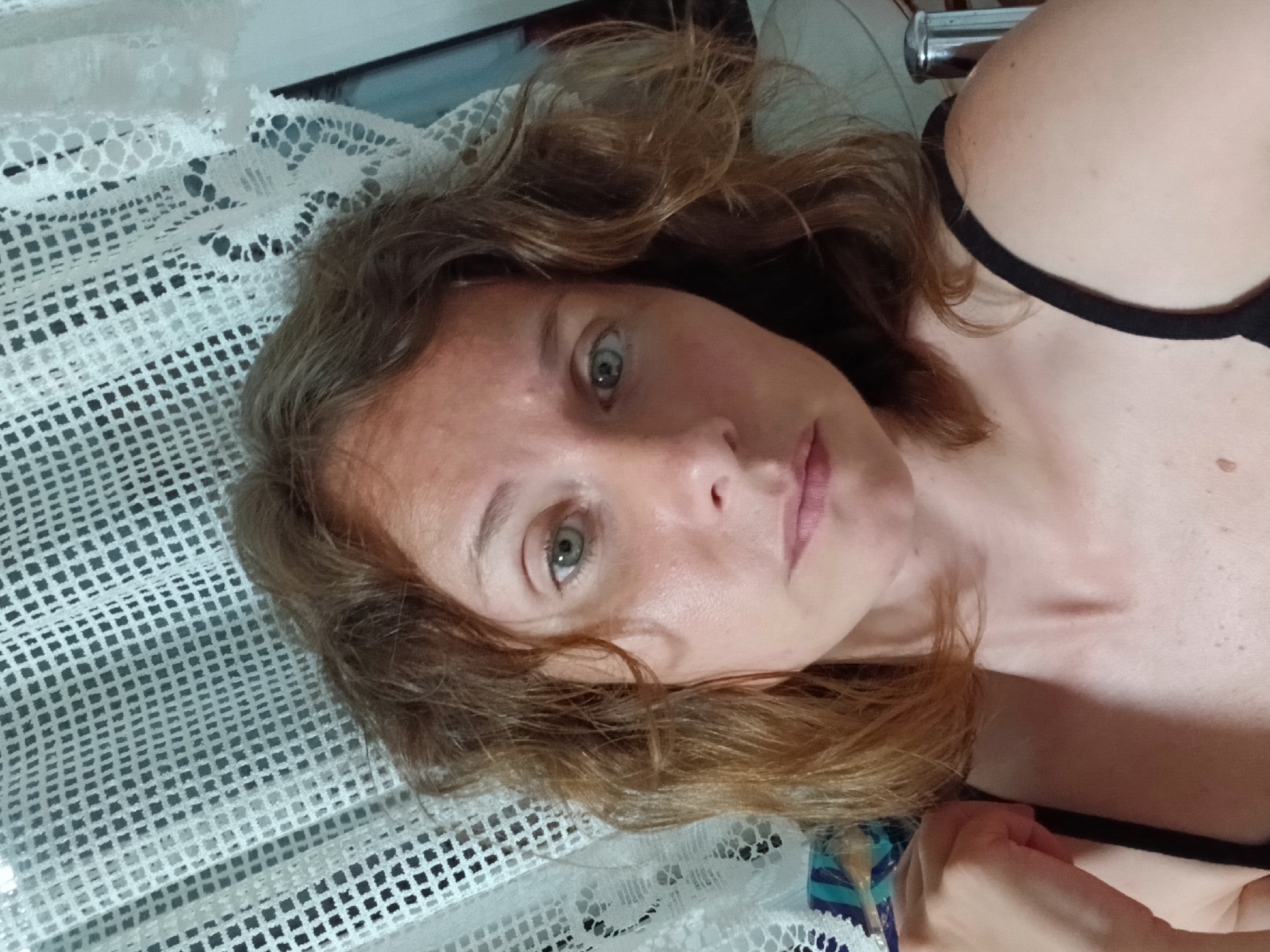 A woman with shoulder-length auburn hair and light-colored eyes is pictured in a close-up, frontal shot. She is looking directly at the camera with a neutral expression. Her skin appears slightly flushed, and there are subtle shadows on her face. She is wearing a black strap garment, and her left arm is visible on the left side of the frame, resting on her shoulder. In the background, a white lace curtain with a square pattern is visible, partially obscuring the view of what appears to be an indoor setting. There are no discernible urban elements suggesting a specific location in Maslova Pristan', Russia.