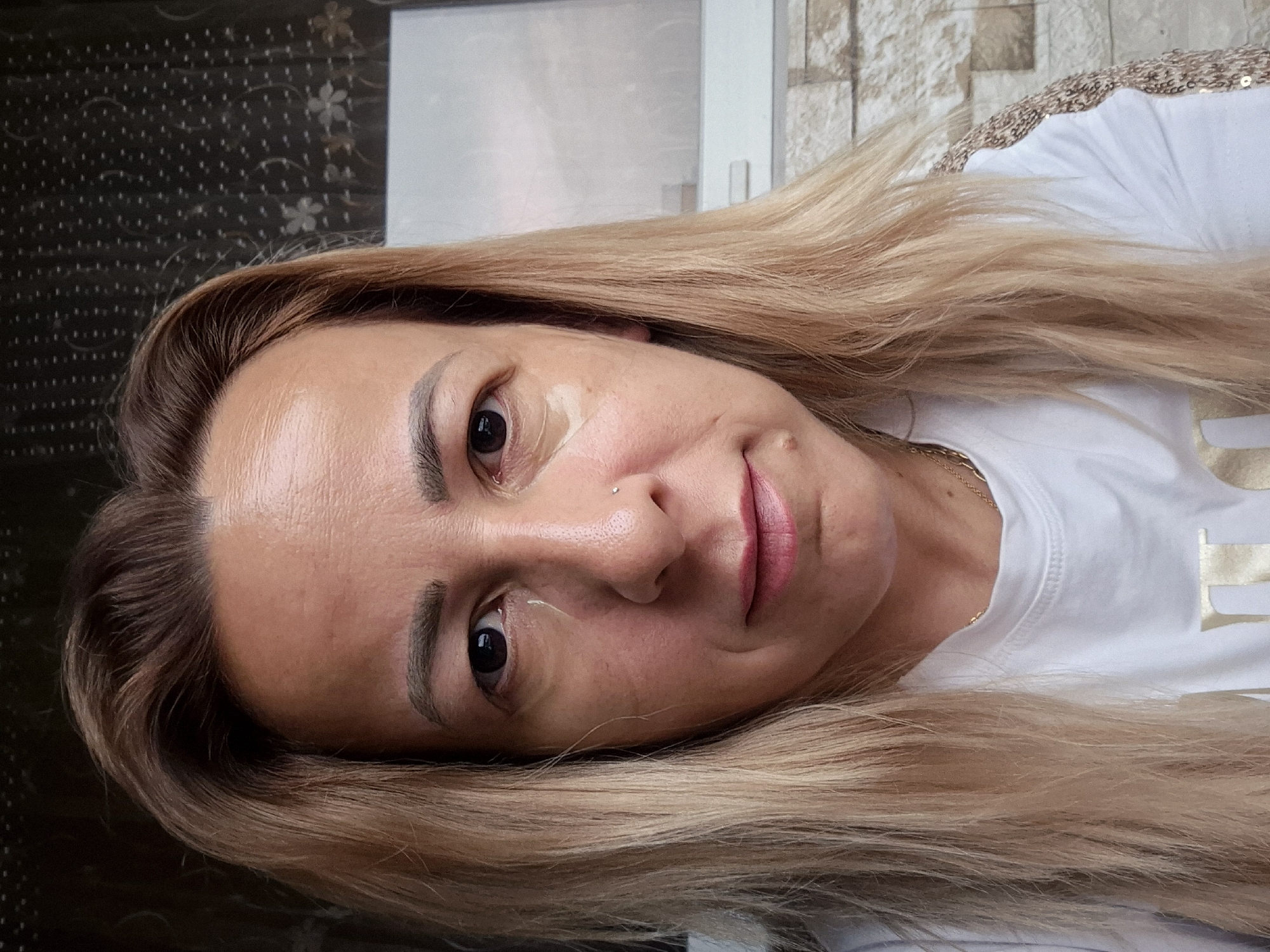 A woman with blonde hair and light skin is taking a selfie. She is wearing a white t-shirt and a gold necklace. There are light beige patches under her eyes. She has a small piercing on her nose. She is looking directly at the camera. The background appears to be a textured wall and a window with a floral pattern. The location is Unknown, Unknown.