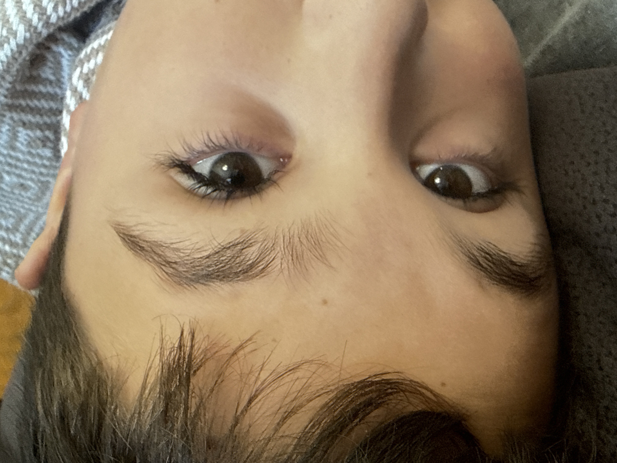 This is a close-up, headshot, eye-level, angled, indoor photograph of a person's face, with the subject lying down. The person has dark hair, brown eyes, and fair skin. There are no visible identifying features or context that would suggest a specific location within the city of Sulitsa, Bulgaria. The environment appears to be a dimly lit indoor setting with a textured blanket or fabric visible in the background. The focus is on the subject's eyes and eyebrows, with a mole visible on the forehead.
