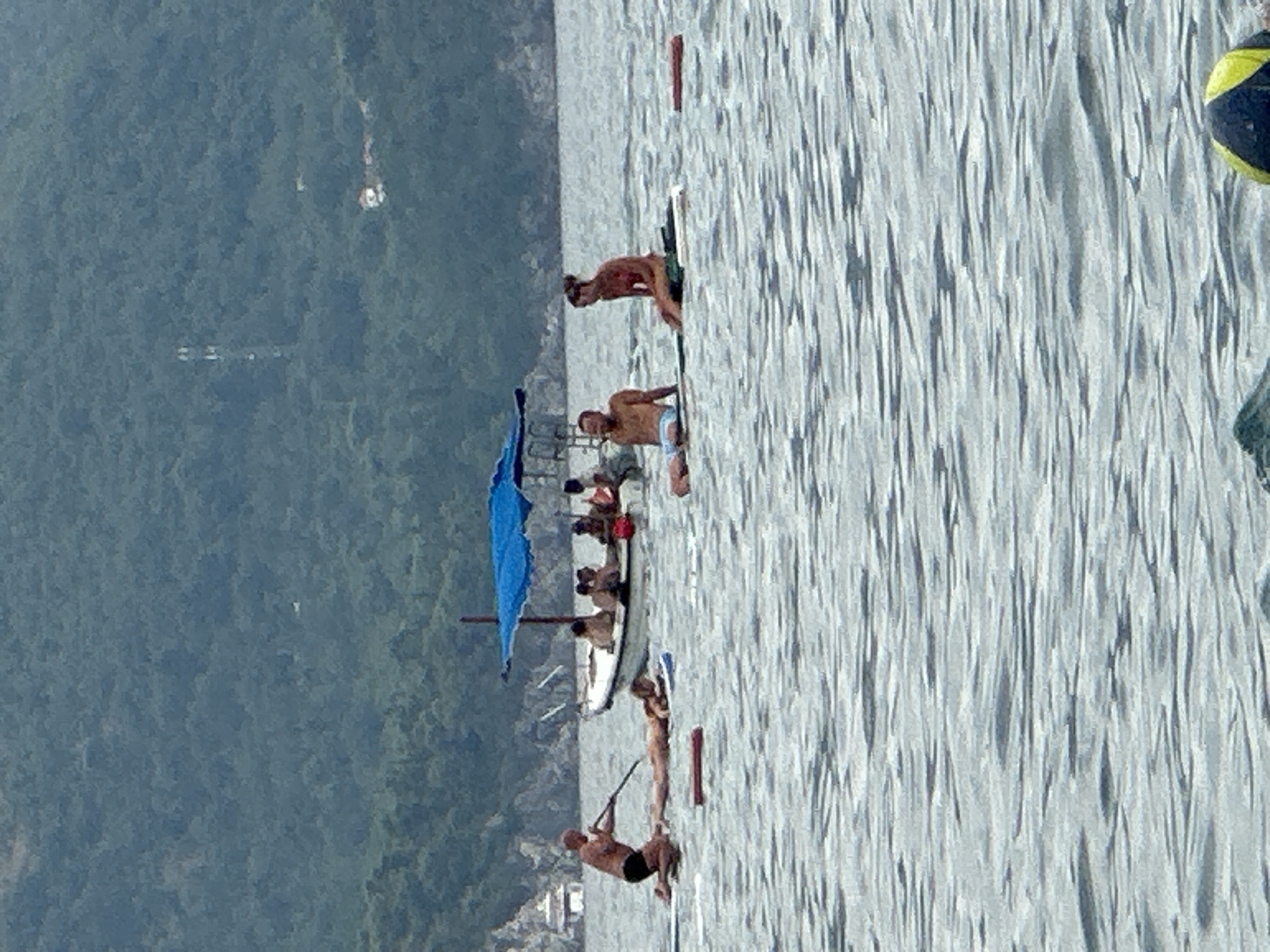 The image shows a view of the coast of Rafailovići, Montenegro, on a cloudy day. In the foreground, several people are seen on paddleboards in the water. One man is kneeling on a paddleboard and appears to be paddling with a pole. Another person is lying down on a paddleboard. Two people are sitting on paddleboards. In the mid-ground, a small boat with several people on board is anchored. The boat has a blue canopy. In the background, lush green hills covered with trees rise from the coastline. A few buildings are visible nestled amongst the trees on the hillside. The water is rippled and appears to be a moderate depth.