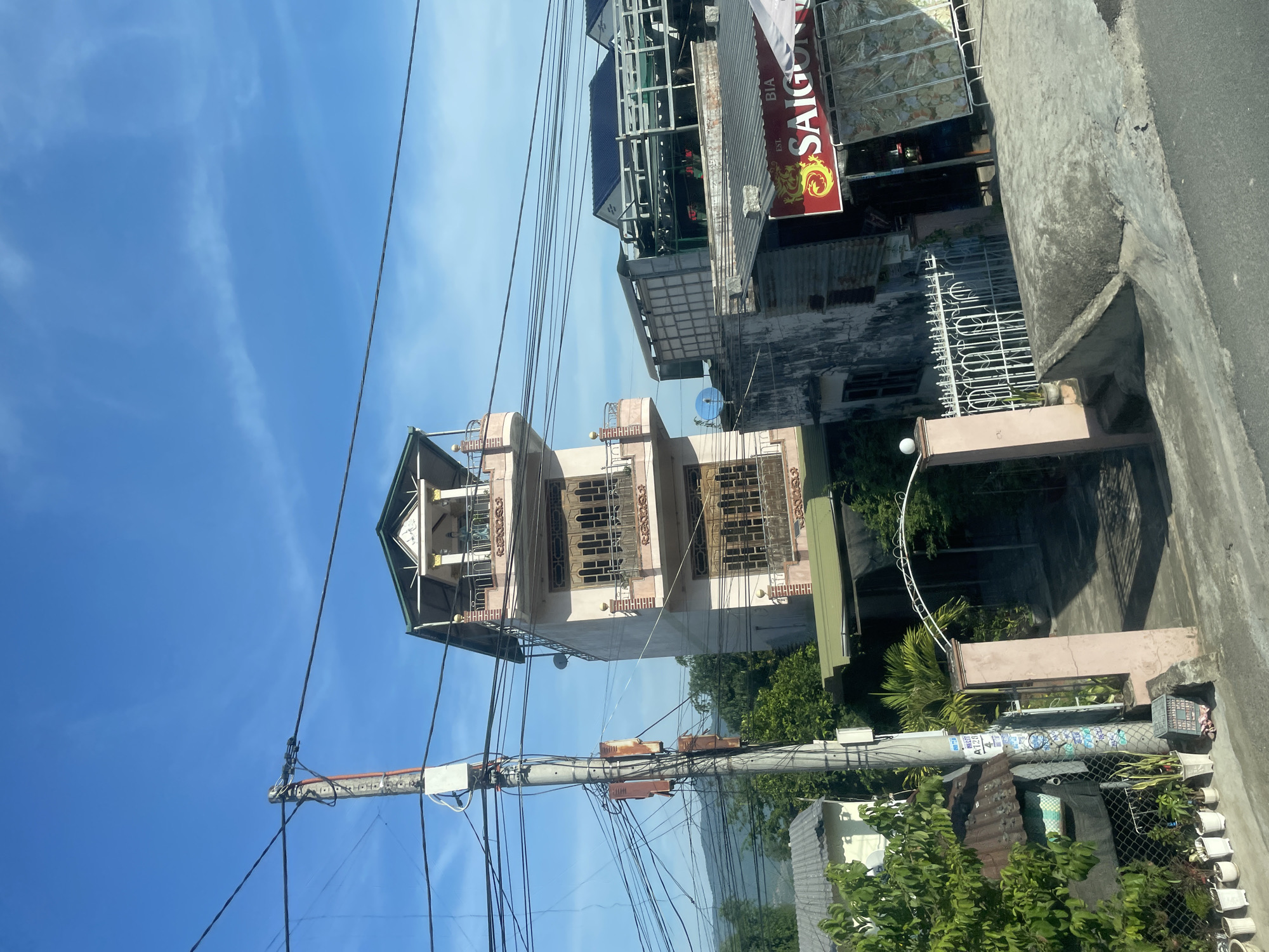 The image depicts a street view in Tay Nha Trang Ward, Vietnam, on a clear day with a bright blue sky. The scene is dominated by a tall, multi-story building with a pink facade and green trim, featuring several balconies with decorative railings and large wooden-framed windows. To the left of this building is a utility pole heavily laden with electrical wires, some of which are rusty. Adjacent to the tall building, on the right, is a lower structure with a corrugated metal roof and a red sign that reads "BIA SAIGON" with a dragon graphic. There are also other buildings and greenery visible, including trees and potted plants. The foreground shows a concrete pathway and a white metal fence. No people are directly visible in the image.