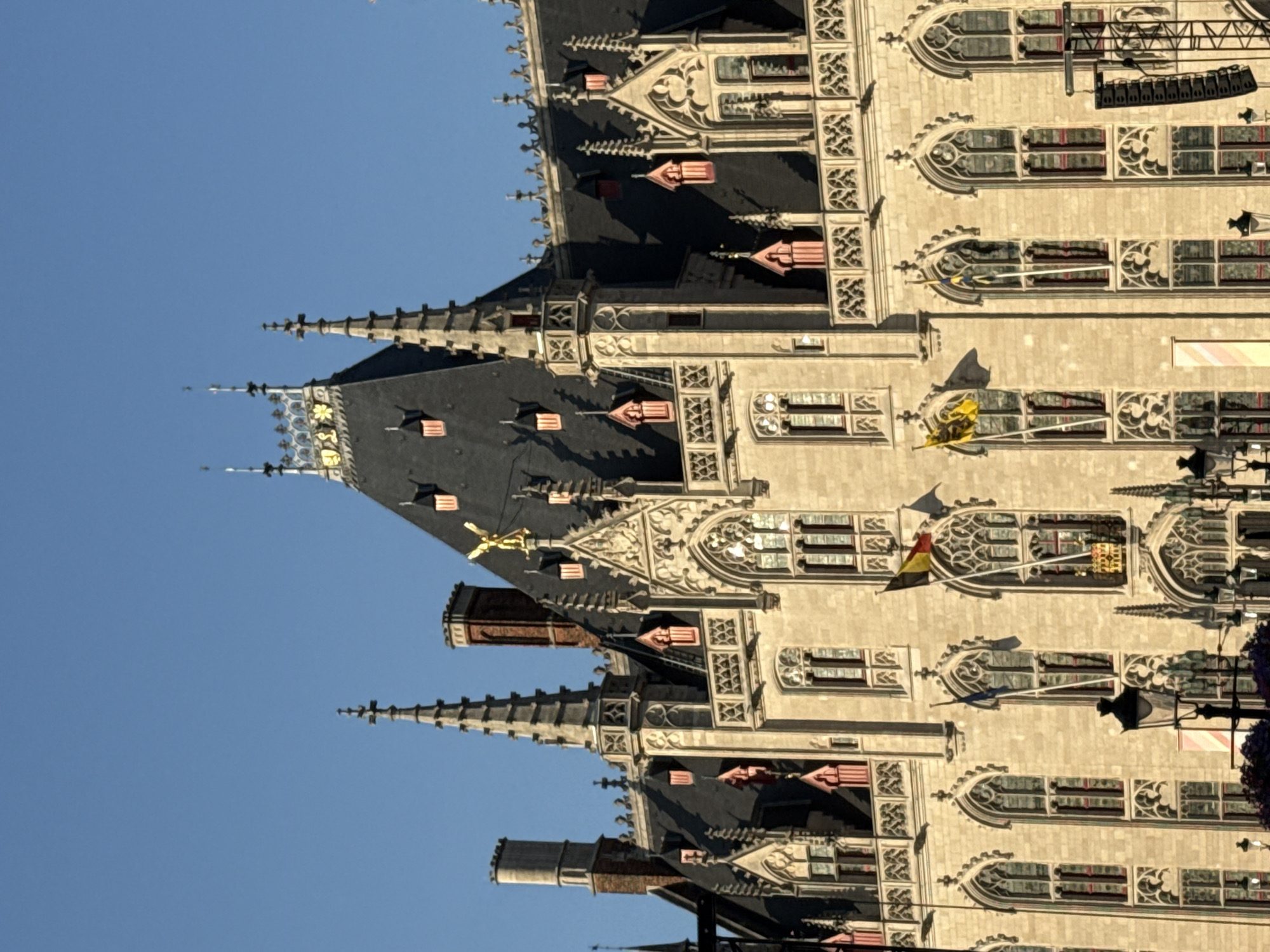 The image captures the ornate facade of the Basilica of the Holy Blood in Bruges, Belgium. The building displays Gothic architecture with intricate stonework, pointed arches, and steep, dark roofs topped with spires. Several flags are visible, including the Belgian flag and what appears to be a regional flag. The sky above is a clear, deep blue, suggesting fair weather. The low angle of the sun casts distinct shadows, highlighting the architectural details. No people are clearly visible in the foreground, but there are hints of activity, such as what could be a temporary stage structure on the right side of the image.
