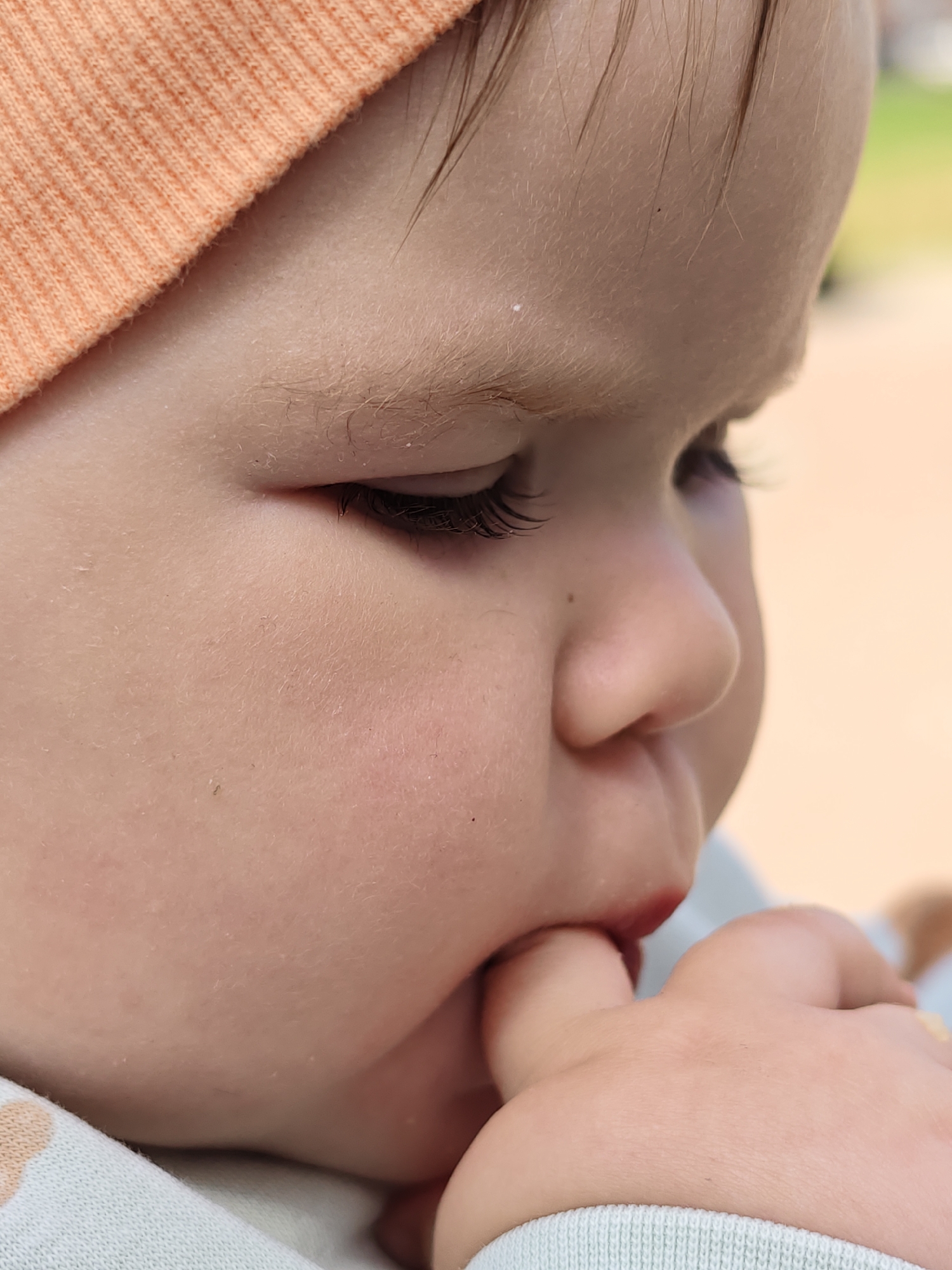 A close-up shows a baby with a light orange knit cap on their head. The baby is fair-skinned with fine hair and delicate eyelashes. The baby's eyes are closed, and their face is turned in profile. The child has their fingers in their mouth. The backdrop appears blurred, suggesting an outdoor environment. Location information is unavailable.
