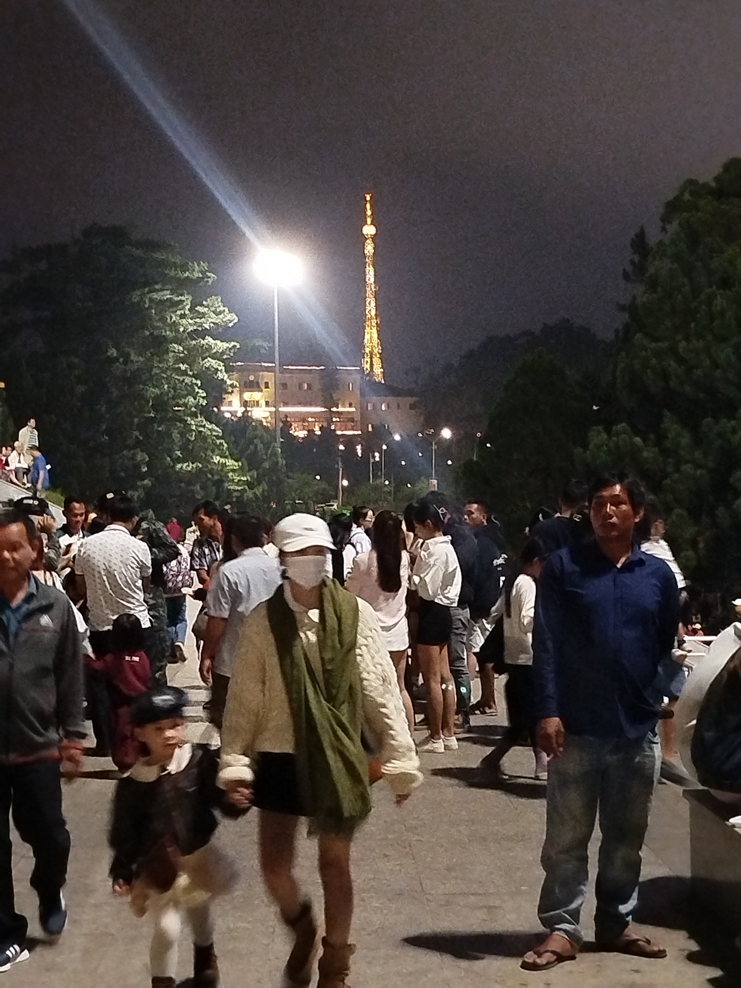 The image depicts a nighttime scene in Da Lat, Vietnam, likely at a popular tourist spot given the presence of a tall, illuminated tower in the background. A large crowd of people are walking on a paved pathway, illuminated by a bright spotlight and the distant tower's lights. The foreground features several individuals, including a woman wearing a white mask and a light-colored jacket, a man in a blue shirt and jeans, and a child in a dark jacket holding hands with the woman. Trees line the pathway, and buildings are visible in the mid-ground. The atmosphere appears to be lively, with many people out and about enjoying the evening.