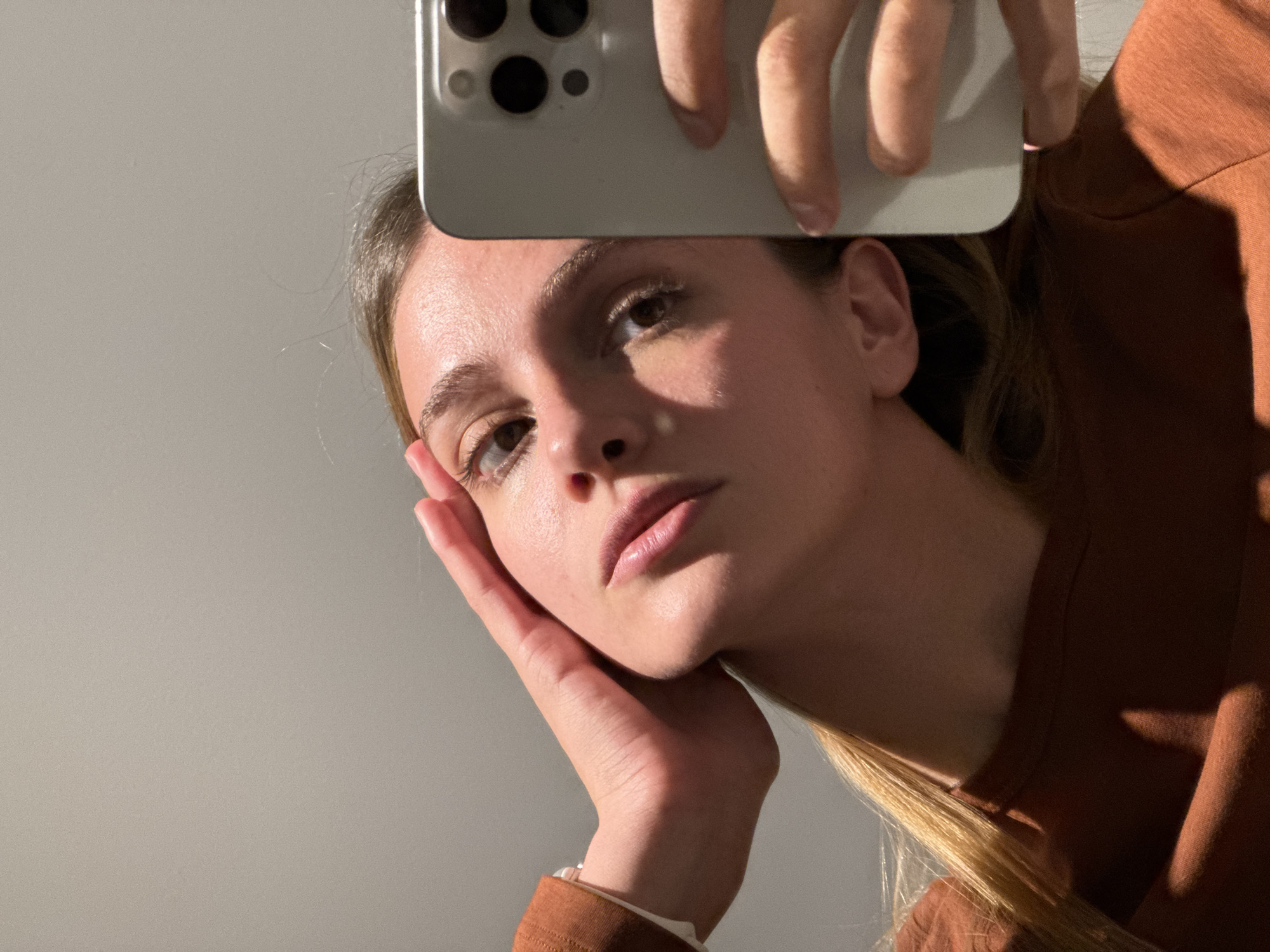 A young woman holds a smartphone above her head, seemingly taking a selfie. The phone is light grey and obscures the top of her forehead. Her left hand supports her head with her fingers angled up towards her face. She wears a long-sleeved shirt that appears brown. Her hair is light blonde and slightly disheveled. The background is a plain, light grey wall. The scene is unlikely to be identifiable as a specific location in Tbilisi, Georgia based on the provided information. There is no indication of an external environment.