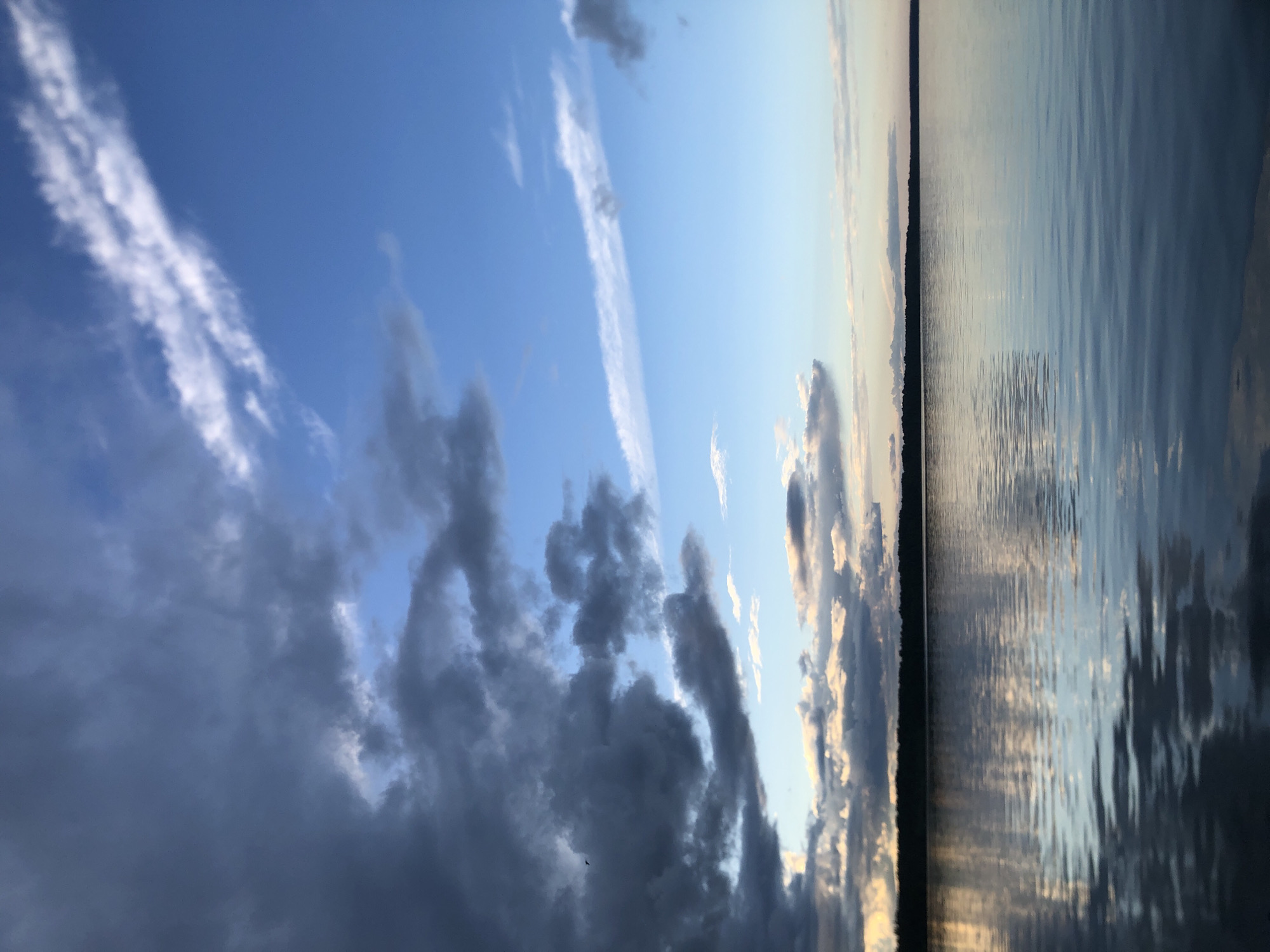 The image captures a serene natural scene, likely a lake at sunset, with the horizon line dividing the sky and water. The sky is a dramatic display of clouds. On the left side, dark, heavy clouds dominate, suggesting impending weather or the remnants of a storm. Interspersed with these are wisps of white, cirrus-like clouds, catching the light. Towards the right, the sky transitions to a clearer, paler blue, with thinner clouds tinged with the warm hues of sunset.
The water on the right reflects the sky, creating a shimmering, rippled surface. The dark line of trees or land is visible along the horizon, separating the sky from the water. The overall impression is one of a quiet, contemplative moment in nature, possibly on the Svirstroy River in Russia, given the context. The lighting indicates it is either early morning or late evening, with the sun low on the horizon. The weather appears to be transitioning, with a mix of dark and light clouds. There is no visible text in the image.