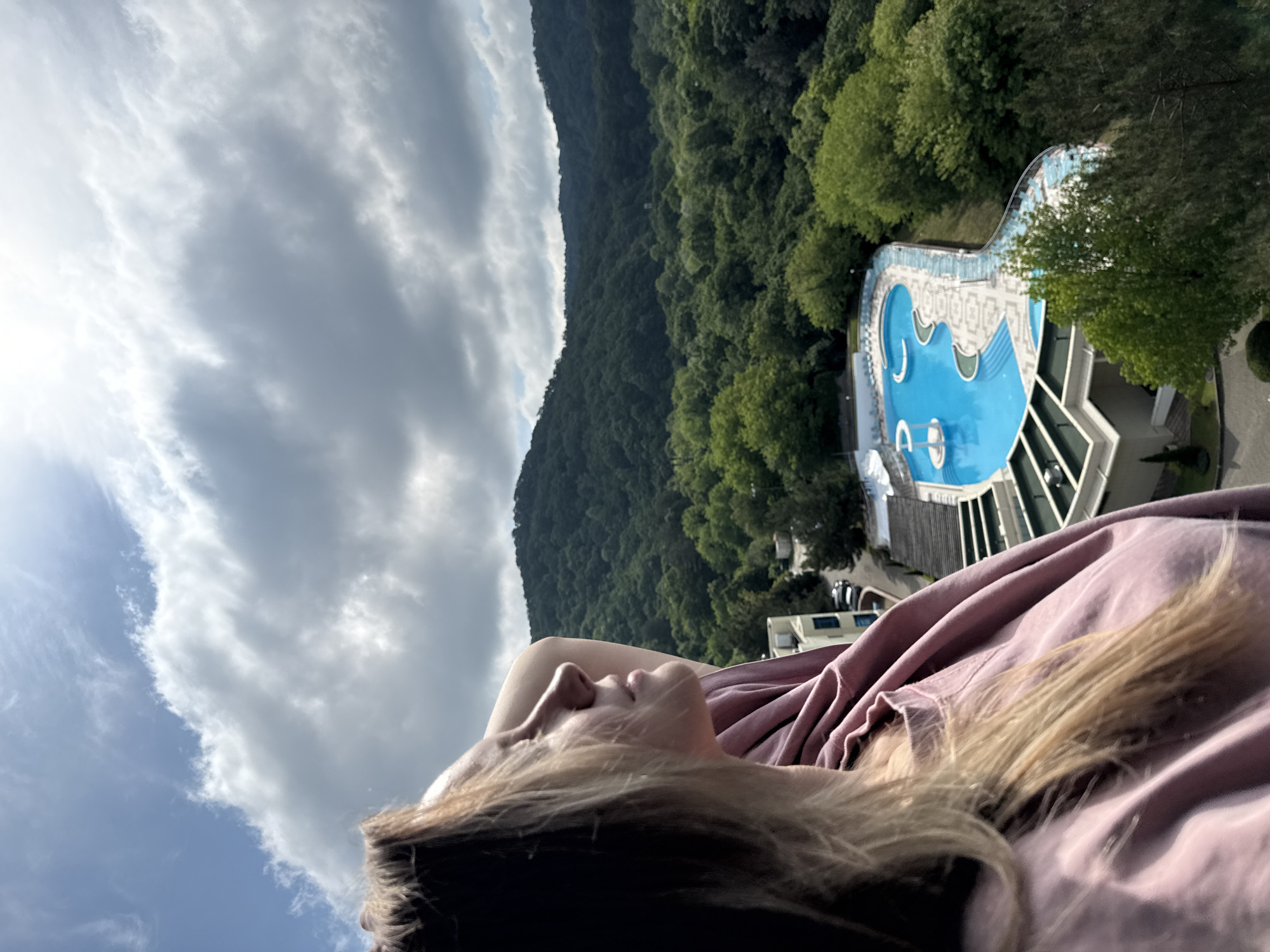 An outdoor, high-angle, medium shot captures a woman in the foreground, her head turned away from the camera and looking up towards a cloudy sky. She has blonde hair and is wearing a pinkish-purple top. Her right arm is raised and resting behind her head. In the middle ground, a large, blue swimming pool with white accents and lounge chairs is visible, surrounded by lush green trees and a building with a light-colored facade. In the background, rolling hills covered in dense forest stretch towards the horizon under a partly cloudy sky. The scene is set in the city of Nebug, Russia.