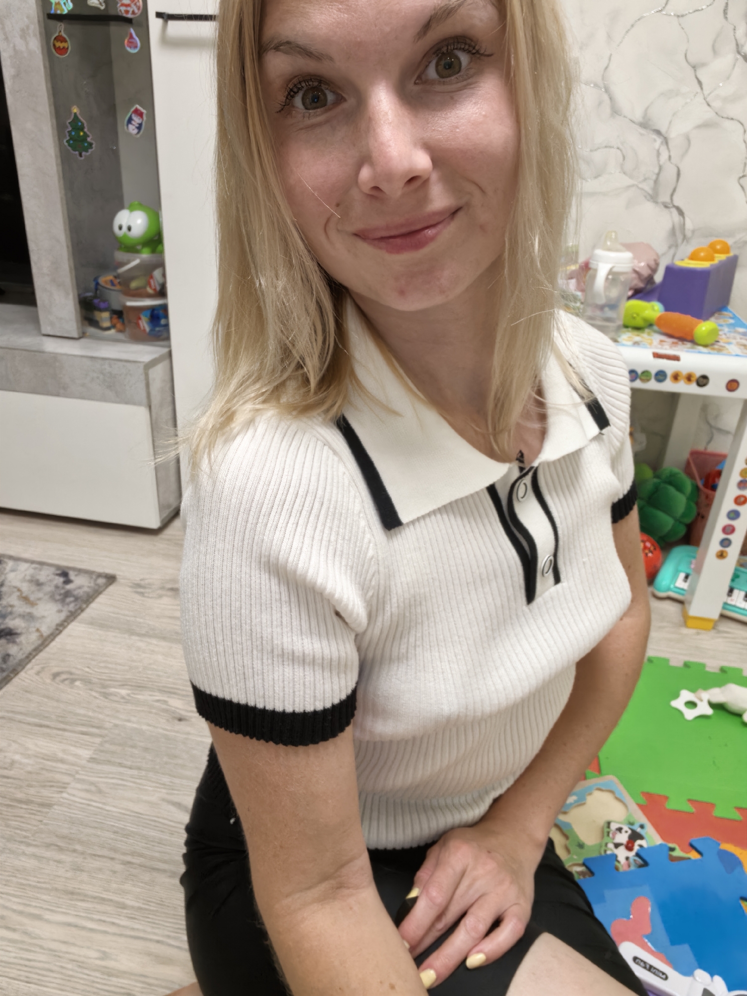 A blonde woman is kneeling on a floor with light-colored wood planks. She is wearing a white, short-sleeved, ribbed polo shirt with black trim on the collar and sleeves, and a black skirt. Her right hand rests on her lap, and her fingernails are painted yellow. Behind her, to the right, is a multi-colored play mat with various children's toys, including a toy keyboard, a plush green toy, and plastic building blocks. To the left of the woman, there is a white cabinet with items on and inside it, including what appear to be stickers on the glass door, one of which is a green character. The background wall has a marbled pattern. This scene does not provide any specific indicators of its location within the city of Sterlitamak, Russia.