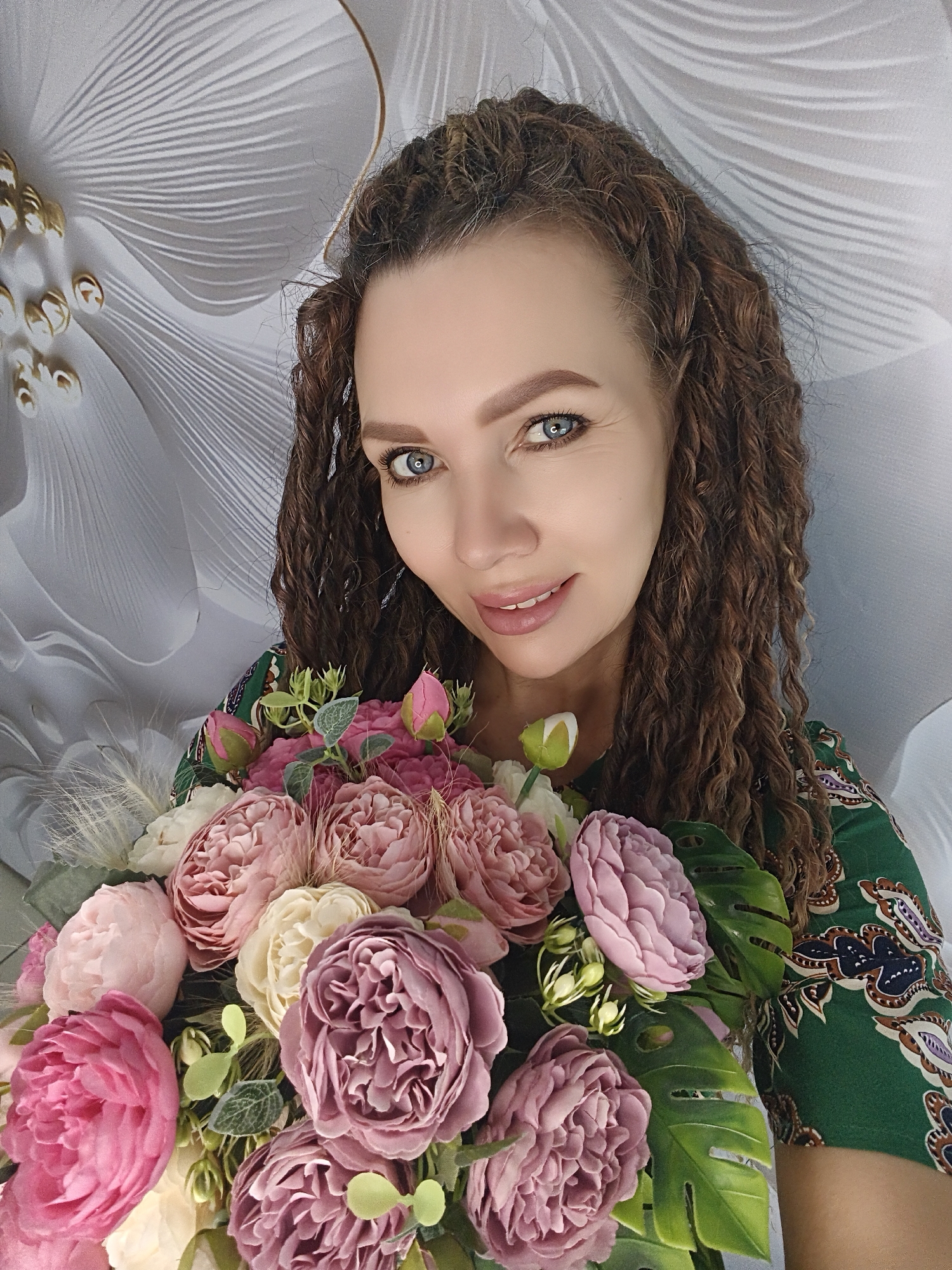 A woman with curly brown hair and blue eyes smiles as she holds a large bouquet of pink, purple, and white flowers with green leaves. She wears a green dress with a paisley pattern. The background is a white wall with a textured, floral design. The location is unknown.