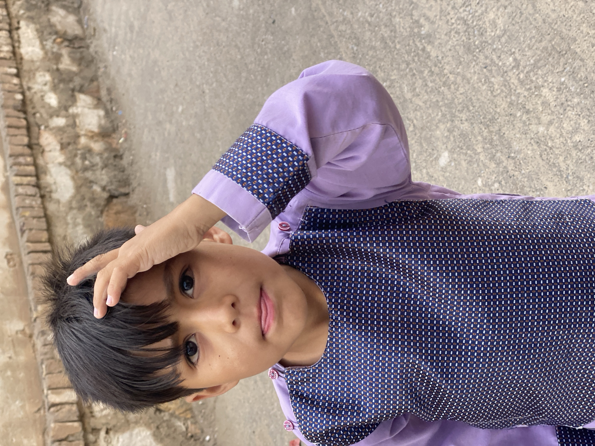 The image depicts a young boy in what appears to be a street or alleyway in Kandahar, Afghanistan. The boy, estimated to be between 5 and 8 years old, has dark, short, unkempt hair and dark eyes. He is wearing a long-sleeved light purple shirt with a dark blue patterned panel covering the chest and upper back. The pattern consists of small, repeating white and orange/brown geometric shapes. The cuffs of his sleeves are folded back, revealing the same patterned fabric. Small, round purple buttons with a distinctive double-line pattern are visible on the shoulder area of his shirt.
The boy's right hand is raised, with fingers splayed, and his palm is resting against his forehead. His gaze is directed forward, towards the viewer, and his mouth is slightly open, with his tongue subtly protruding.
The background consists of a weathered, light brown brick wall, showing signs of age and possibly disrepair, with visible mortar lines. The ground beneath the boy is a light grey, rough, concrete or asphalt surface, appearing somewhat uneven with minor debris. The lighting is consistent with outdoor daylight.