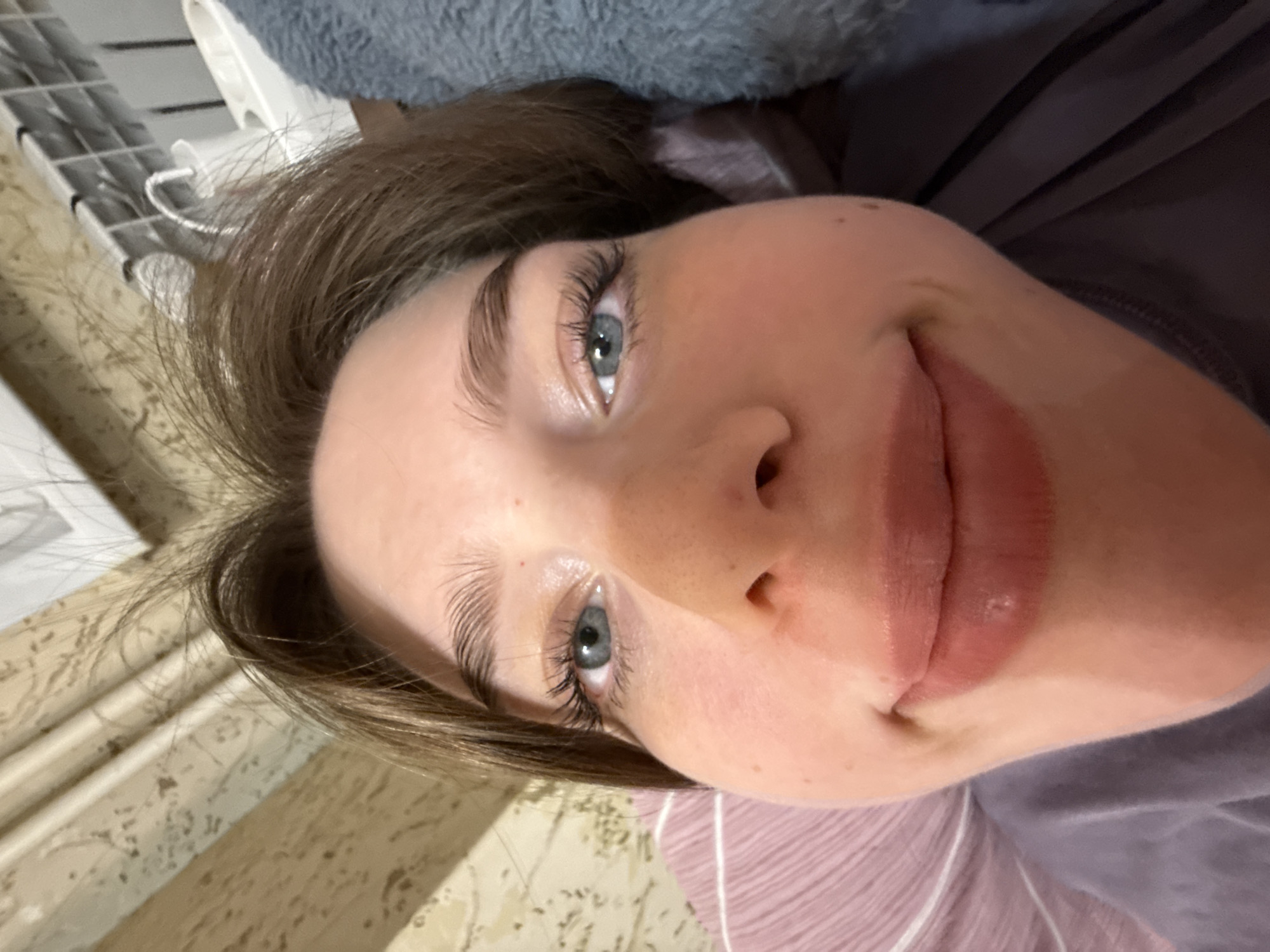A close-up, tilted selfie depicts a woman with light skin, light brown hair, and blue eyes. She is smiling slightly, looking directly at the camera. The background includes a wall with light-colored patterned wallpaper on the left, a white object (possibly part of a window or air conditioning unit) near the top left, and what appears to be clothing or bedding in shades of gray and purple surrounding her face. There is no information to indicate the location is Krasnoyarsk, Russia.
