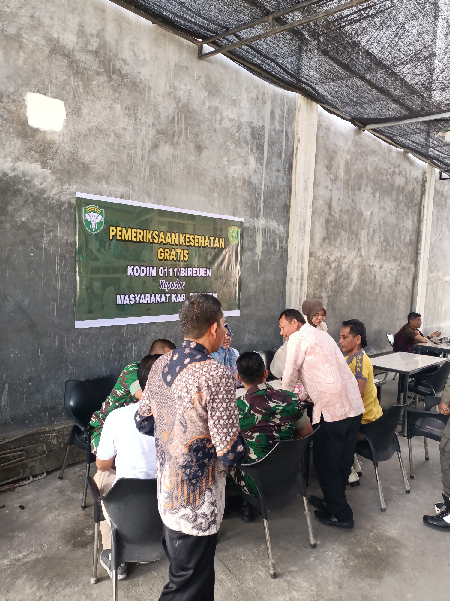 In the city of Cotgadong, Indonesia, a free health check-up event is taking place, evidenced by a banner in the background reading "PEMERIKSAAN KESEHATAN GRATIS KODIM 0111/BIREUEN Kepada: MASYARAKAT KAB." The scene unfolds in an open-air shaded area with a concrete wall and floor. Several individuals are gathered around tables and chairs, some in military uniforms, others in civilian attire. One person in the foreground wears a patterned shirt, facing away from the camera. To the left, individuals in camouflage uniforms are seated, suggesting they are participants or staff. In the background, more people are seated at tables, some appearing to engage in conversations or receive services. The atmosphere suggests a community event focused on health services.
