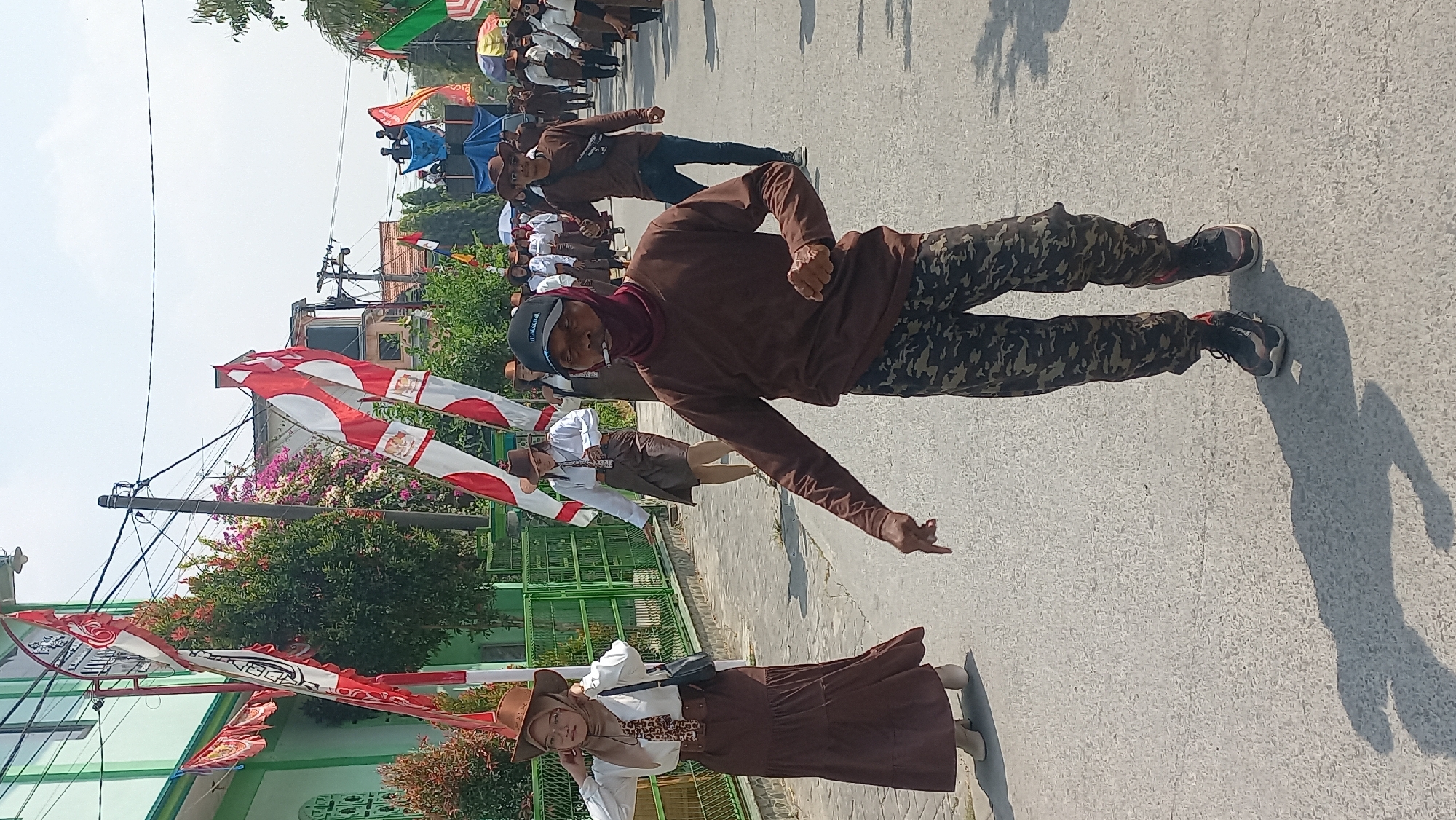 In Kedungmegarih, Indonesia, a procession is underway on a paved street under a clear sky. A group of people, some dressed in traditional brown attire with hats, are walking. Two women in the foreground wear brown long skirts and white blouses, with one adjusting her headscarf. A man in a brown long-sleeved shirt and camouflage pants, with a bandana covering his lower face, walks forward with his right arm extended and index finger pointing. He is smoking. Several Indonesian flags with red and white stripes are being carried by participants. In the background, more people are visible, some on a raised platform with speakers, and buildings line the street, one with green paneling. Trees and foliage are present on either side of the road.