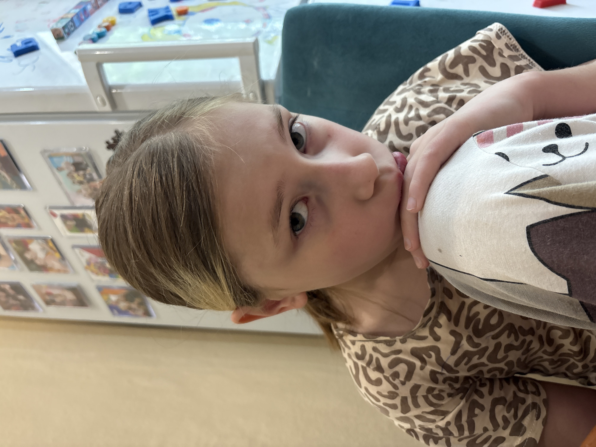 A young girl with blonde and brown hair is the subject of the image. She is wearing a tan and brown patterned shirt and patterned pants. She is positioned in front of a blue upholstered chair. A white refrigerator is visible to her left, adorned with multiple rectangular magnets. On top of the refrigerator is a clear plastic container containing small toys of various colors. The photograph was not taken in the city of Veliky Novgorod, Russia.