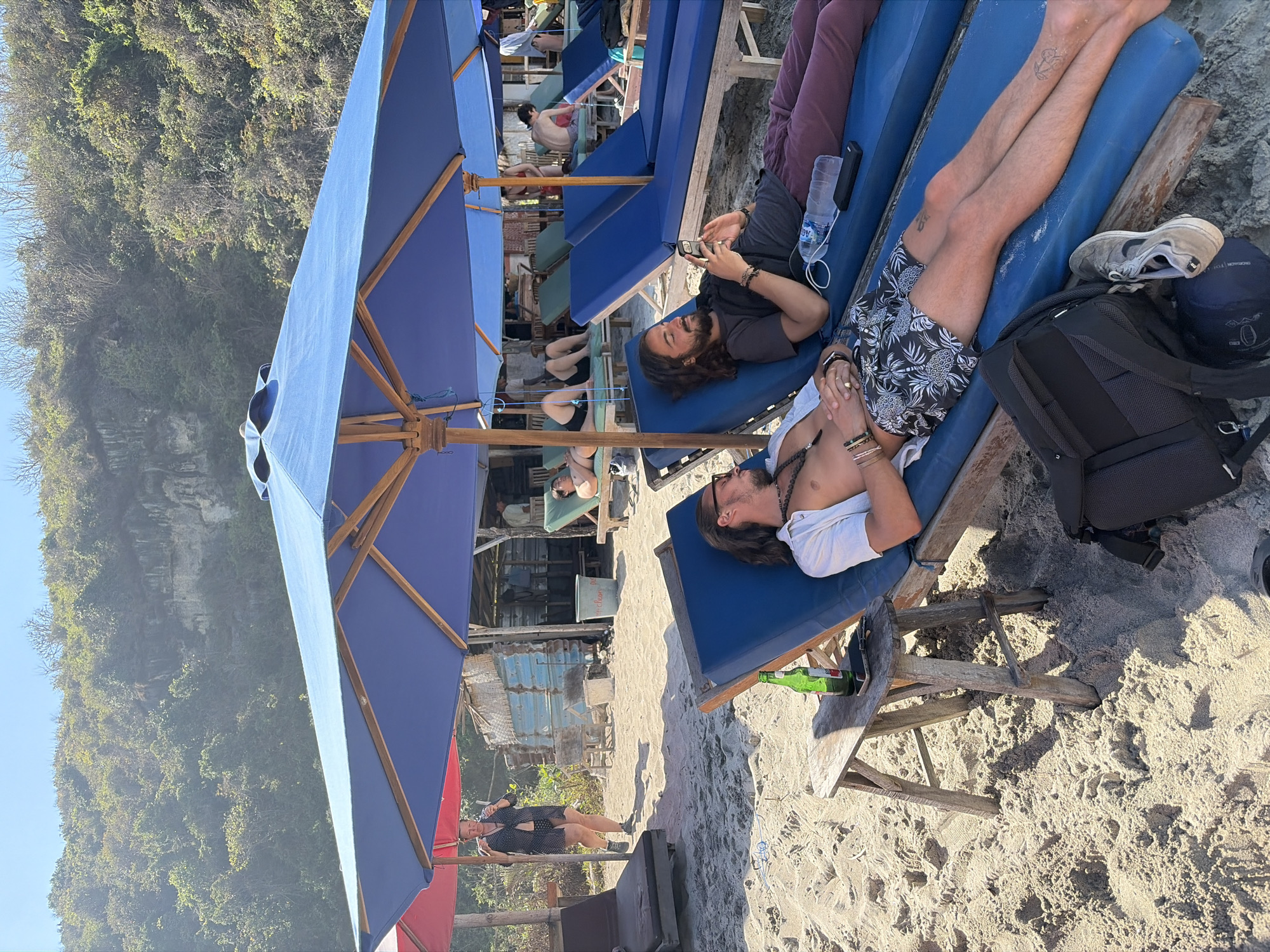 This image depicts a beach scene in Nusa Penida, Indonesia, with people relaxing under large blue umbrellas. In the foreground, two men are lying on lounge chairs. The man on the left is wearing a white open shirt and patterned shorts, with his eyes closed. Next to him is a wooden side table with a bottle of beer and a mobile phone. The man on the right is wearing a dark t-shirt and maroon pants, looking at his phone. A backpack and a pair of sneakers are visible on the sand near the lounge chairs.
In the background, other individuals are seen on lounge chairs under umbrellas. A woman in a dark polka-dot outfit is walking in the sand near a small structure made of corrugated metal and wood. The environment is sandy, with lush green vegetation and a rocky cliff face visible in the distance, indicative of Nusa Penida's coastal landscape. The overall atmosphere is one of leisure and relaxation on a sunny day.
