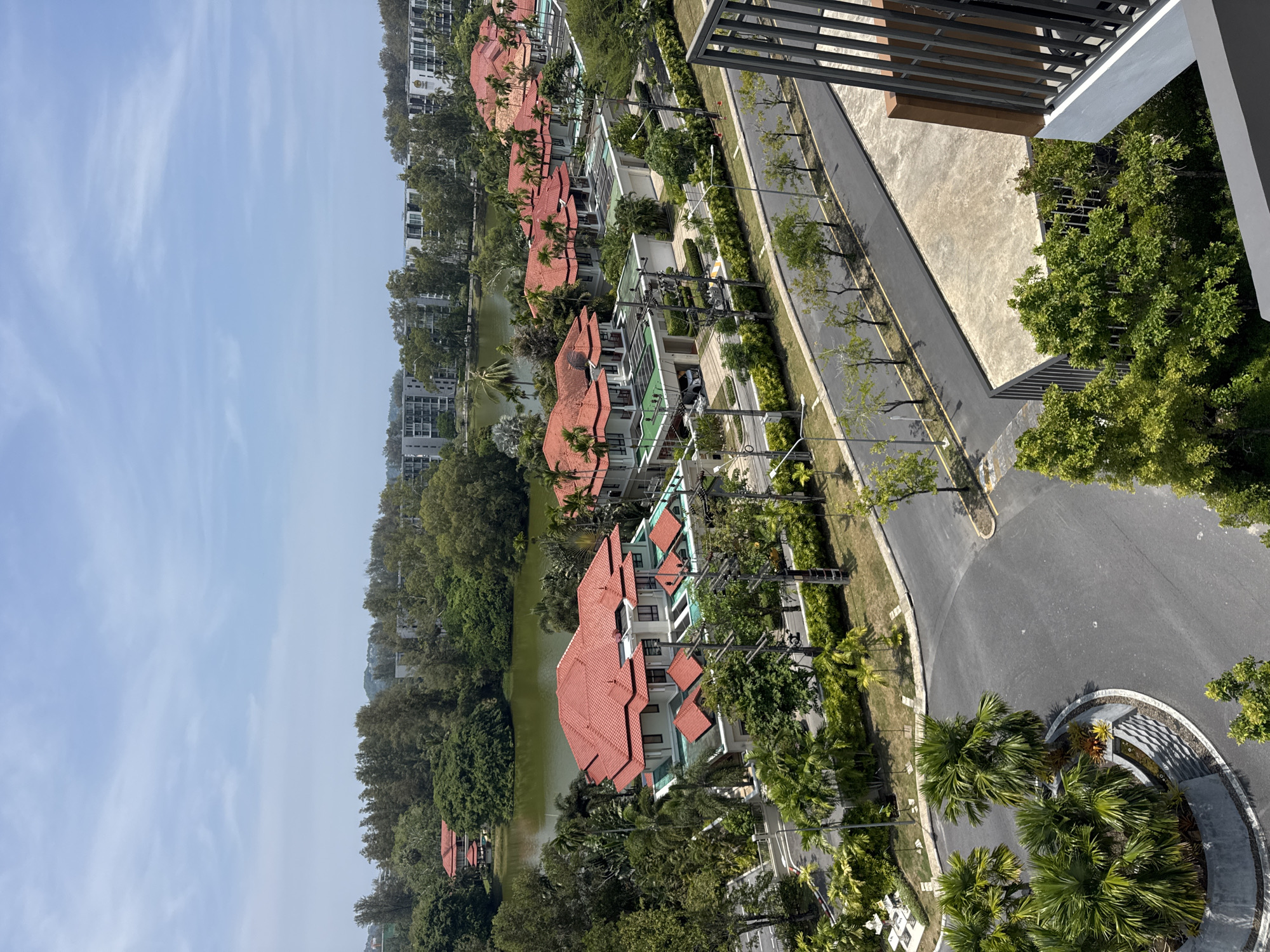 An aerial view shows houses with red tiled roofs lining a lake. A road runs parallel to the houses, with palm trees and other trees planted along the roadside. The sky is light blue with scattered clouds. The image appears to be taken from a high-rise building, with a balcony visible in the foreground. The location is Choeng Thale, Thailand.