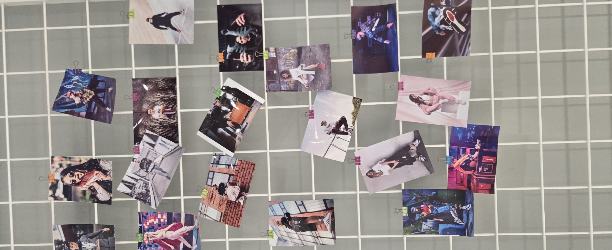 This media file captures a grid board, likely a white wire mesh, affixed against a light grey wall. Numerous small, printed photographs, resembling instant camera prints, are attached to the grid using various colored binder clips (pink, green, orange, purple, and blue).
The subjects of the photographs are predominantly young adults, shown in a variety of poses and settings. Many images feature individuals posing for fashion or lifestyle shots, dressed in modern casual wear and sportswear, including sneakers, plaid shirts, and tracksuits. The scenes are diverse, depicting individuals dancing, lounging, or standing against urban structures, natural backdrops, or indoor environments. The overall impression is a collection of personal moments, fashion spreads, or promotional imagery.
The photographs are arranged in a dynamic, seemingly casual manner, clipped at different angles and sometimes overlapping, creating an informal yet artistic display. There is no singular event taking place, but rather a compilation of individual moments. The lighting appears even, suggesting an indoor setting. While the location context is provided as Budva, Montenegro, no specific visual cues within the images or on the grid board explicitly identify this location. No discernible text is visible on the clips or the grid, and any text within the photographs is too small to read.