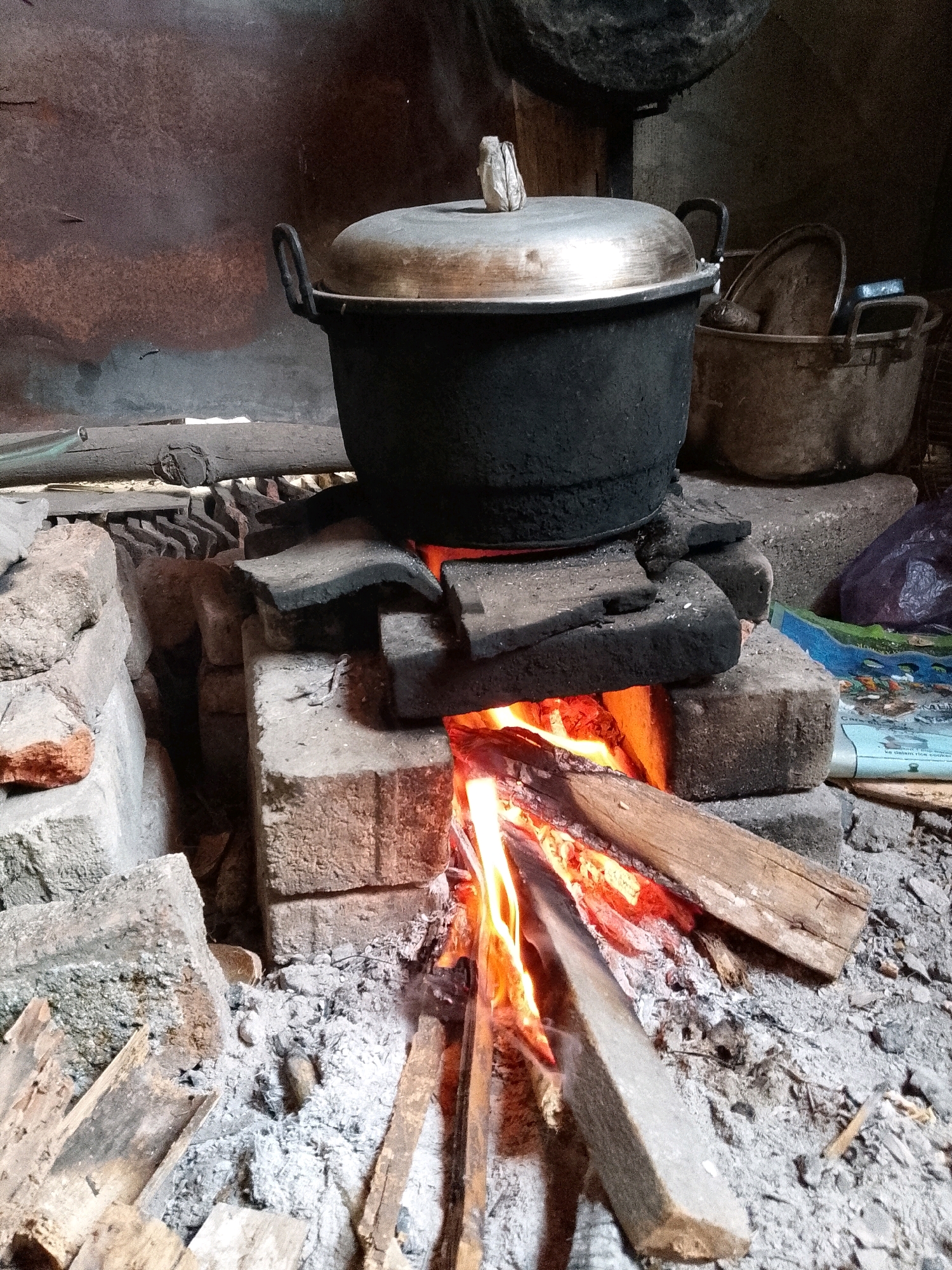 The image depicts an outdoor kitchen scene in Wadang, Indonesia, focusing on a traditional wood-fired stove. A large, dark pot with a metallic lid is positioned over the fire, resting on a structure made of bricks and flat stones. Flames from burning wood are visible beneath the pot, illuminating the surrounding area. The ground is covered with ash and scattered pieces of wood, indicating recent or ongoing use of the stove. In the background, more cooking utensils and a plastic bag are present. The overall atmosphere suggests a rustic and functional cooking environment.