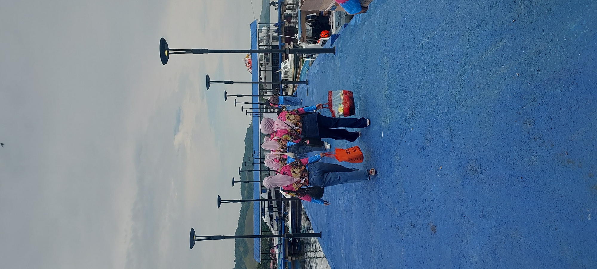 The image depicts a group of people walking on a bright blue pier in Asei Besar, Indonesia. In the foreground, two women are walking away from the camera, both wearing pink headscarves and vibrant, patterned shirts. The woman on the left is carrying an orange shopping bag, while the woman on the right holds a red and white striped bag filled with yellow items. Behind them, several other individuals, also wearing similar attire, follow along the pier. The pier is lined with modern black lampposts. In the background, a body of water is visible, with various boats docked, including a larger white ferry. Hills covered in green vegetation rise in the distance under an overcast sky. The overall scene suggests a community outing or excursion at a waterfront location.