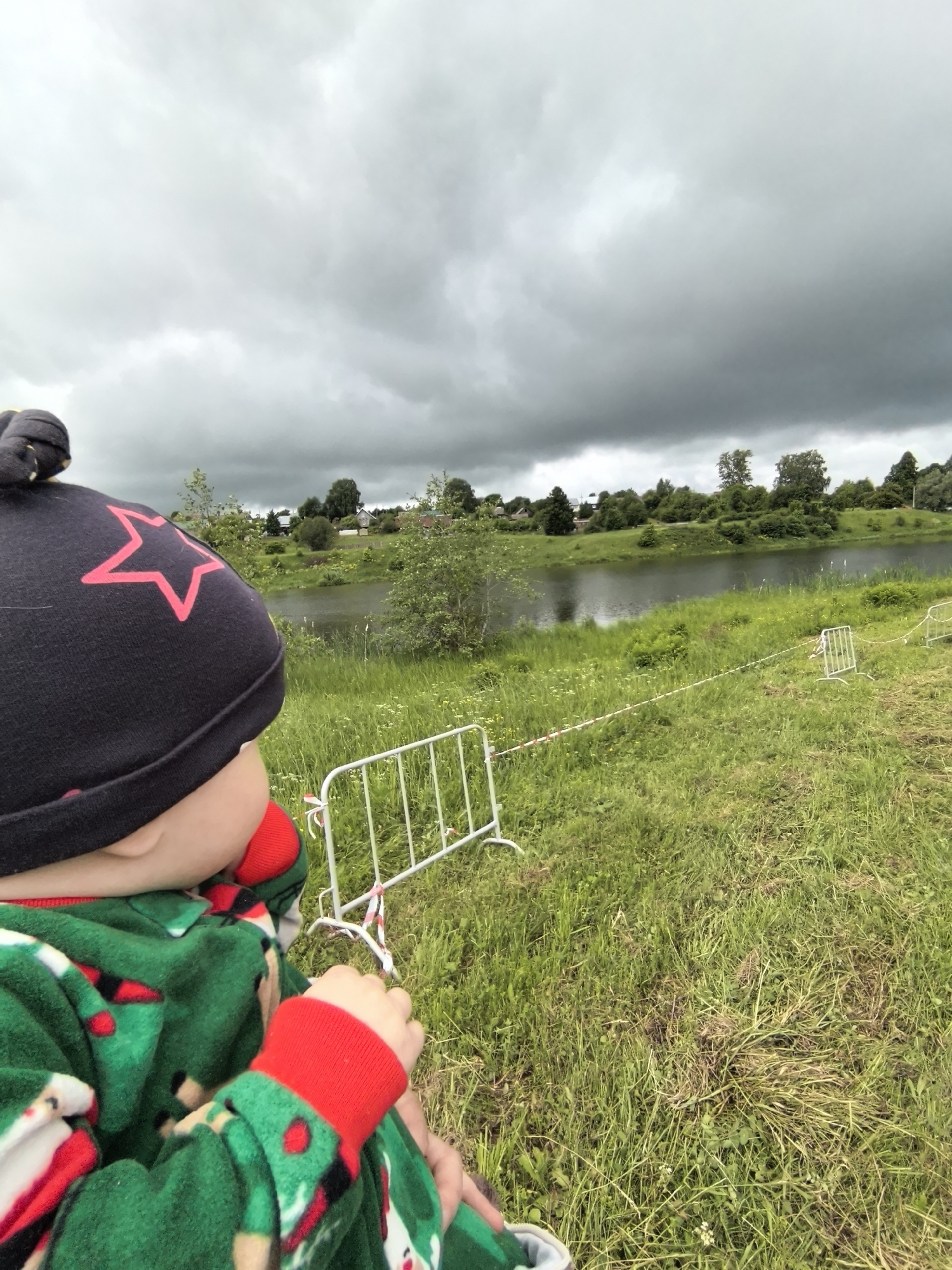 A child, seen from the back of their head and upper torso, is positioned in the foreground, looking leftward towards a landscape under a dark, overcast sky. The child wears a dark beanie with a prominent red star outline and a green, patterned onesie with red cuffs. In the midground, a body of water, likely a river, reflects the grey sky. The banks are covered in lush green grass, interspersed with various small plants and wildflowers. White, portable metal barriers are visible on the grassy bank, with red and white striped tape or ribbon strung between them, delineating an area. Beyond the water, a treeline obscures part of the view, with several houses or buildings partially visible in the distant background, suggesting a small settlement. The ground in the immediate foreground is covered in green grass with some matted patches. The scene is located in the city of Unknown, Unknown.