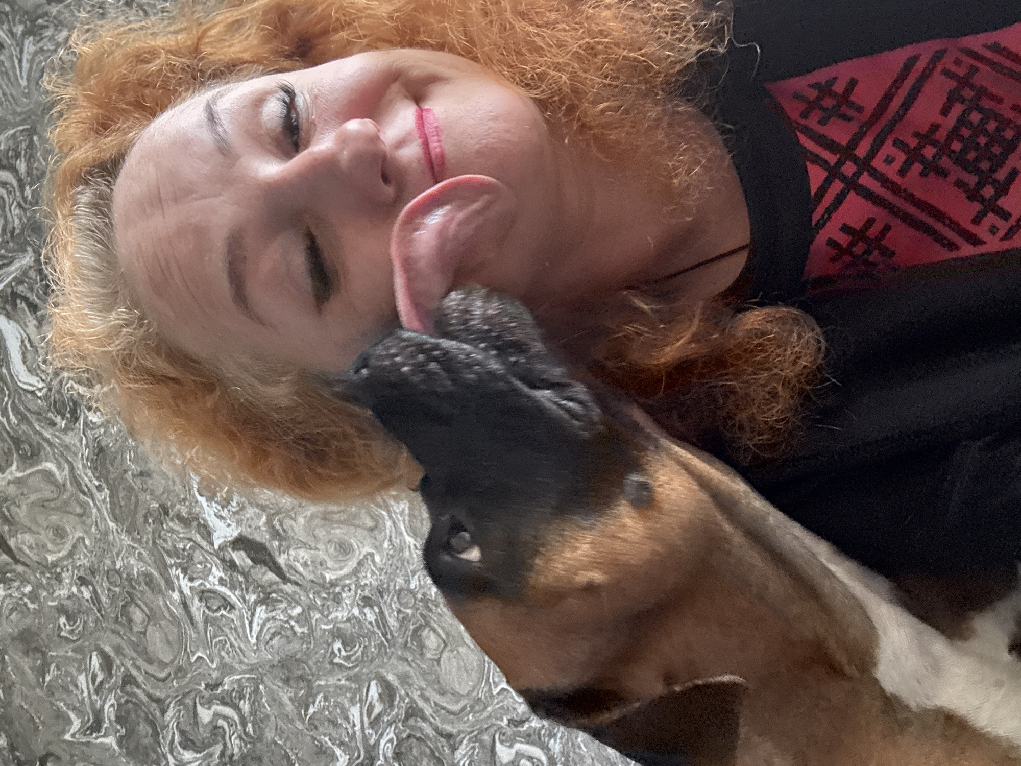An overhead shot captures a warm moment between a woman and a dog, set indoors in Chaikovsky, Russia. The woman, with vibrant red curly hair, is lying down with her eyes closed and a wide smile on her face, indicating joy and affection. She is wearing a black garment with a section of red fabric featuring a distinct black geometric, possibly embroidered, pattern.
The dog, a medium-sized breed with short, brown and black fur and some white markings, is actively licking the woman's cheek, its tongue fully extended. The interaction suggests a strong bond and playful affection between the two. The woman's head rests on a surface with a swirling, marbled pattern in shades of grey and white, which could be bedding or a textured floor.
The lighting appears to be ambient indoor light. The close-up nature of the shot emphasizes the expressions of contentment on the woman's face and the dog's enthusiastic display of affection. No visible text is present in the image.