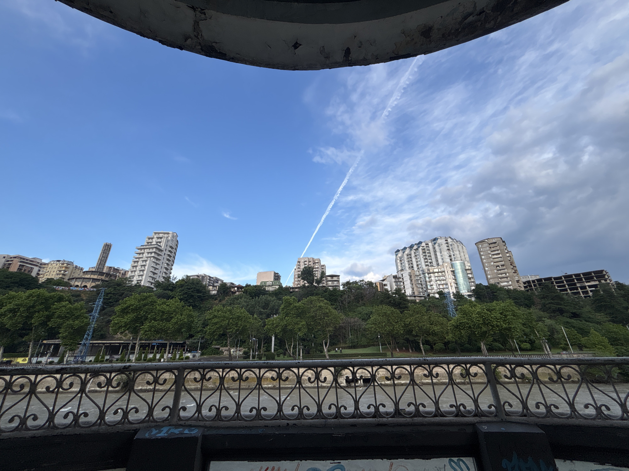 The image captures a view from a balcony or elevated walkway overlooking the Mtkvari River in Tbilisi, Georgia. In the foreground, a decorative metal railing with heart-shaped scrollwork separates the viewpoint from the river below, where the water appears somewhat murky. Behind the railing, a treelined embankment slopes upwards, dotted with buildings of various architectural styles. Prominent among these are several modern apartment buildings, some appearing as high-rises. A construction crane is visible amongst the trees on the left side of the embankment, suggesting ongoing development. The sky above is a vibrant blue, streaked with wispy white clouds and a distinct contrail from an aircraft. The overall scene depicts an urban landscape integrated with green spaces and the natural element of the river.