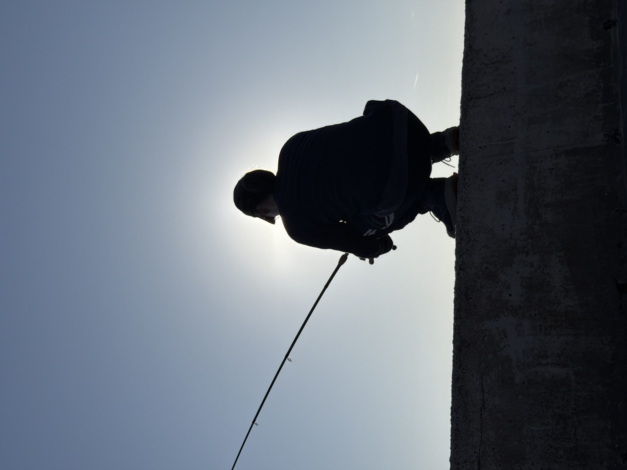 The image captures a solitary figure silhouetted against a bright, clear sky, engaged in fishing. The scene is shot from a low angle, emphasizing the person and the vastness of the sky.
The main subject is a person, visible from the waist up, rendered as a dark silhouette due to intense backlighting from the sun positioned directly behind them. They are wearing what appears to be a hooded garment or jacket. The person is crouched or leaning forward on a rugged, dark grey concrete wall or pier, which occupies the right side of the frame. They are actively holding a fishing rod, which extends diagonally downwards and to the left, with its tip pointing towards the bottom left corner of the image. The rod has visible eyelets along its length.
The background consists entirely of a bright, light blue sky, indicating daytime with clear weather. A faint white contrail from an aircraft is visible in the upper right area of the sky. The strong sunlight creates a vibrant halo around the person's silhouette.
The activity depicted is fishing, with the individual's posture suggesting concentration and engagement with their rod. The location context indicates this activity is taking place in Petrovac centar, Montenegro, likely on a pier or seawall. No text is visible within the image, and the person's emotions are not discernible due to the silhouette.