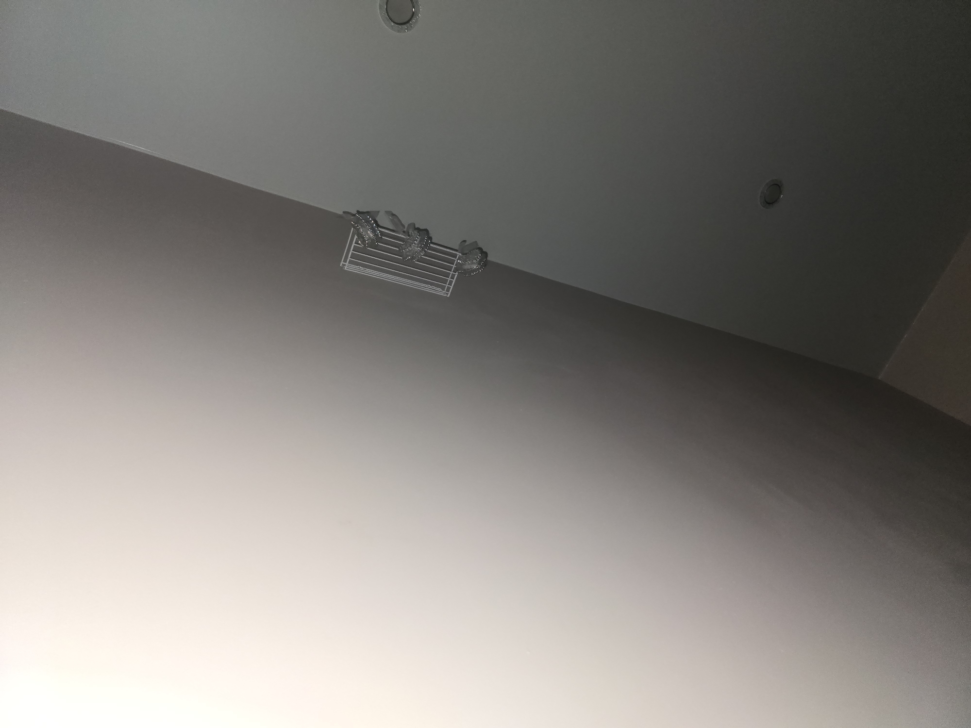 The image shows a ceiling with a white shelf mounted on it. There are decorative items on the shelf that appear to be silver or metallic. The ceiling is painted in a light color and transitions to a darker shade in some areas, suggesting a change in light or a design element. There are at least two recessed light fixtures visible on the ceiling. The overall environment appears to be indoors, possibly a bathroom or a well-lit room. The image does not provide any specific information about the city of Ulan-Ude, Russia, or its location within it.