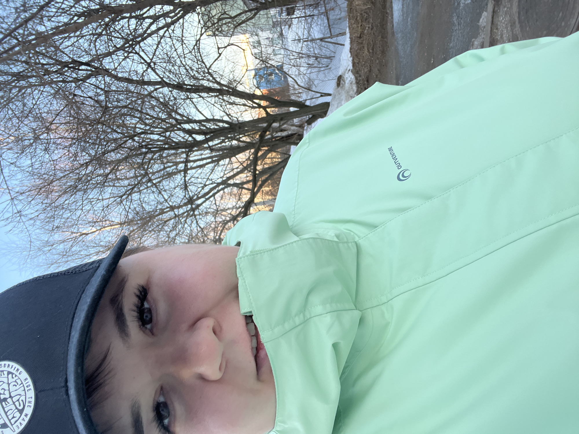 The image displays a close-up selfie of a person, identified as female, taken outdoors. The individual is looking directly into the camera. She is wearing a black baseball cap with a white circular logo on the front, partially showing text such as "LIVE THE WAVE RIDE THE" and "SURFING." She has dark hair and wears a mint green hooded jacket with the collar pulled up, obscuring her chin and mouth, though her upper lip and teeth are visible as she bites the collar. The jacket features a dark grey "OUTVENTURE" logo on the left chest. Her eyes have prominent eyelashes.
The background reveals a clear blue sky above a sparse urban or semi-urban landscape. Numerous deciduous trees with bare branches are visible, suggesting a late winter or early spring setting. The ground contains patches of melting snow mixed with exposed brown earth and dirt. In the mid-ground, a wire fence is partially discernible behind snowdrifts and bare bushes. Distant, indistinct buildings, including a prominent light blue structure, are visible through the trees. The lighting is natural, with a warm hue on the horizon, indicative of sunrise or sunset. The specific location within Chaikovsky, Russia, cannot be determined from the visual evidence.