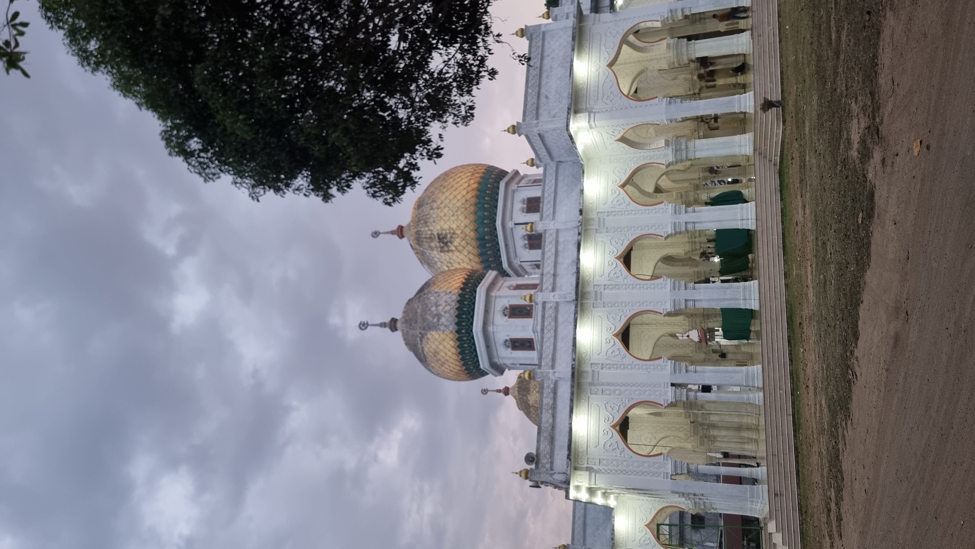 The image depicts the exterior of a mosque in Meunasahlhok, Indonesia, likely during dusk or a cloudy day. The mosque features white walls adorned with intricate Islamic geometric patterns above a series of arched colonnades. Two prominent golden domes, capped with crescent moons, rise above the main structure, with a smaller golden dome visible behind them. The colonnade is illuminated by artificial lights. In the background, through the arches, green drapes and what appear to be prayer mats are visible, suggesting the mosque is operational. A large tree with dark green foliage is visible in the upper right corner, partially obscuring the sky. The ground in front of the mosque is a mix of dirt and patchy grass. Several individuals are present, including one person standing to the right of the mosque entrance, and others are faintly visible within the colonnade. The sky is overcast with grey clouds.