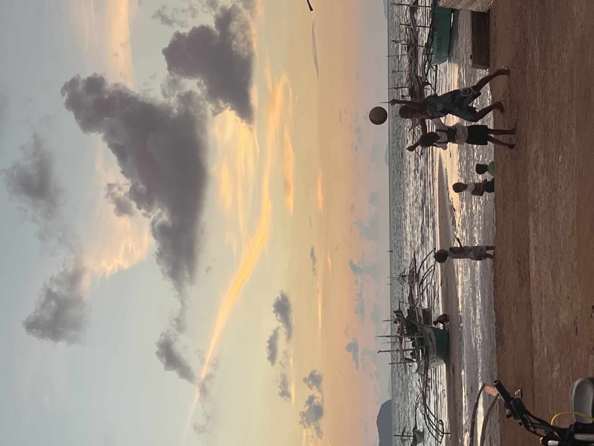 The image depicts a sunset scene at a beach in Cuyo, Philippines. In the foreground, a sandy beach is visible, with the handlebars of a motorcycle on the left. Further back, the ocean meets the shore, with several traditional outrigger boats partially submerged in shallow water. A group of children are playing on the beach and in the water. Two children are actively engaged in a game of volleyball, with one jumping to hit the ball in the air. Other children are observing or playing nearby. The sky is filled with dramatic clouds in shades of orange, yellow, and gray, indicating the setting sun. The overall atmosphere is one of a peaceful evening by the sea.