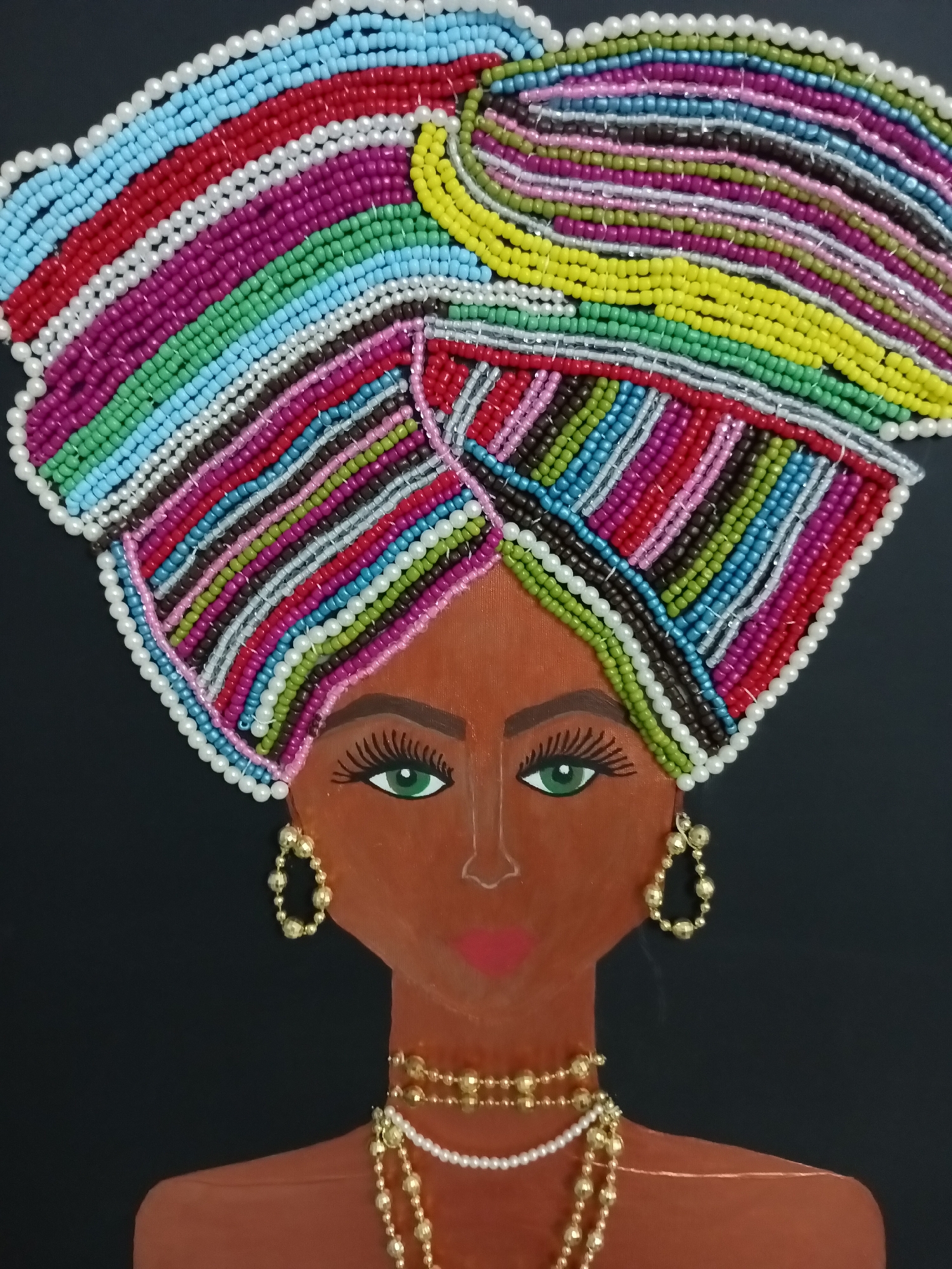 This is a close-up, eye-level, vertical shot of a beaded artwork depicting a woman's head and shoulders against a plain black background. The artwork features a prominent, vibrantly colored turban made of numerous small beads in stripes of blue, red, white, purple, green, yellow, and other hues. A white beaded trim outlines the turban. The woman's face is painted in a brown tone with green eyes, dark eyelashes, and red lips. She is adorned with gold hoop earrings and multiple strands of gold and pearl necklaces. The artwork has no discernible connection to Alanya, Turkey.