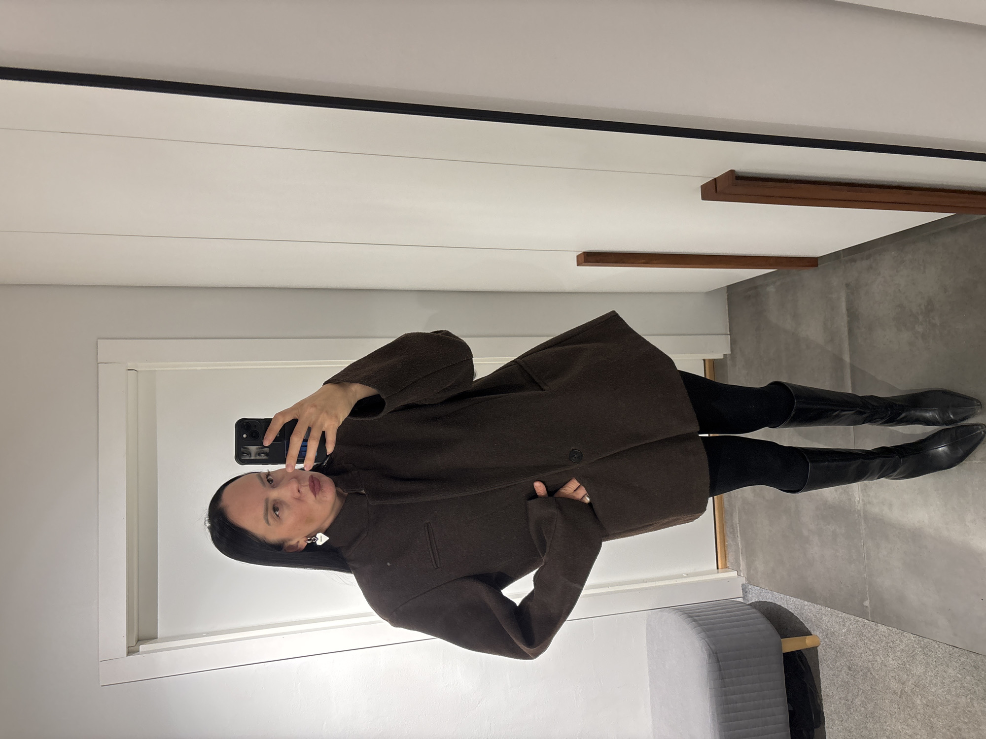 A woman is captured in a mirror selfie, rotated 90 degrees clockwise from its natural orientation. She stands indoors, looking directly at her smartphone which she holds up to take the picture, reflecting her eyes on the screen. She has dark hair and a neutral expression, wearing a dark brown, possibly wool, knee-length coat that appears oversized or loosely fitted. Underneath, she wears black leggings or tights and black knee-high boots. She has prominent heart-shaped earrings.
The setting appears to be a modern interior, possibly a dressing room or a contemporary home in Chaikovsky, Russia. The background features a white wall with horizontal paneling, reminiscent of shiplap. A thin black horizontal line runs along the top of the wall. Further back, two wooden rectangular accents are mounted on the wall, with one appearing as a shelf or decorative beam. The floor is a light gray, concrete-like material. In the lower right corner, a gray upholstered bench or stool is partially visible. The overall aesthetic is clean and minimalist, with subtle textures from the wall and floor materials. The lighting is even, suggesting an indoor artificial light source. No legible text is visible in the image.