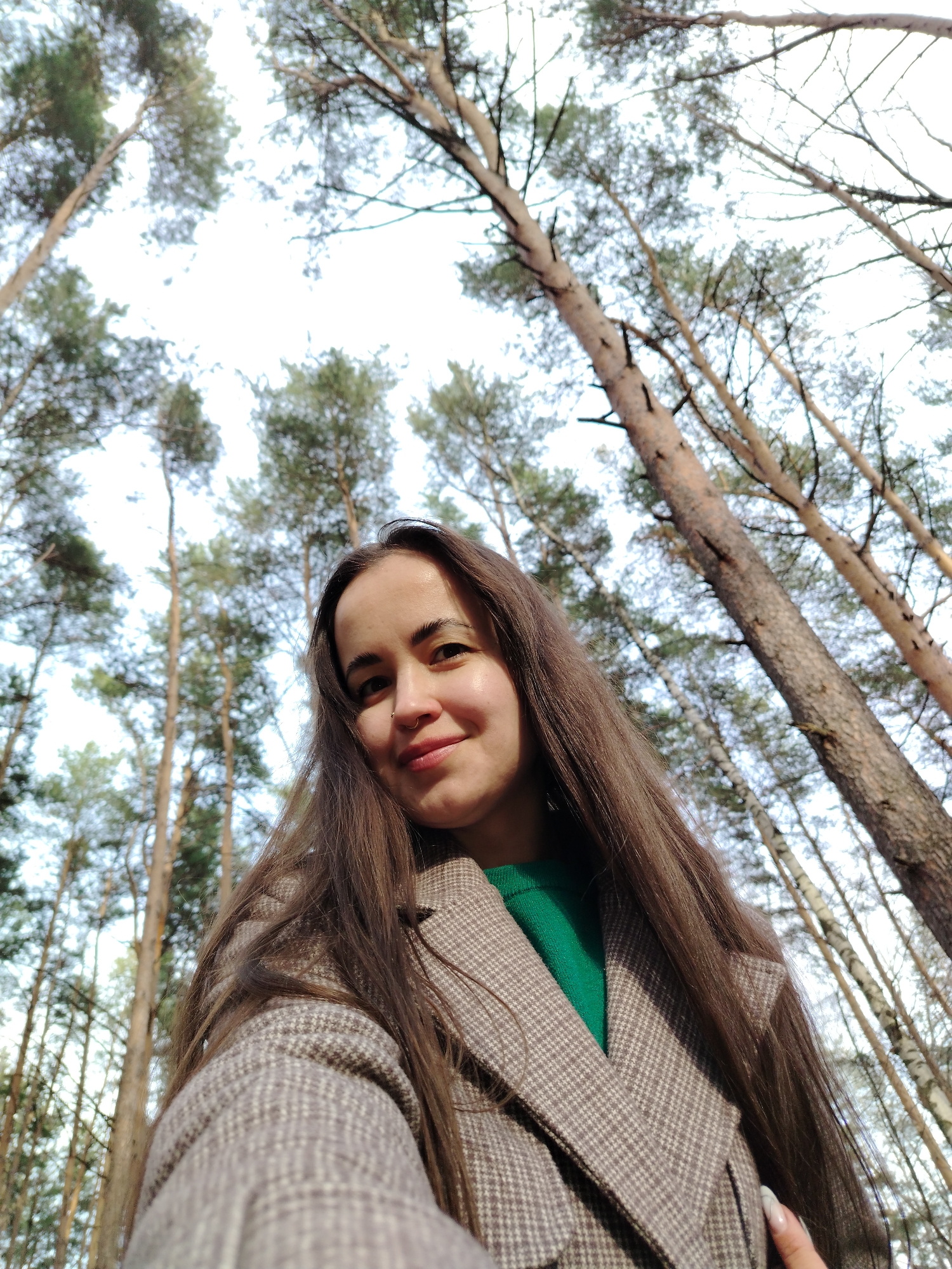 A young woman is taking a selfie in a forest of tall pine trees with a sparse undergrowth of birch saplings. The location is assumed to be near the city of Chaikovsky, Russia. The woman has long brown hair and is wearing a green sweater under a gray checkered coat. The photo is taken from a low angle, looking up at the woman and the trees. The sky is visible through the trees and appears to be overcast.