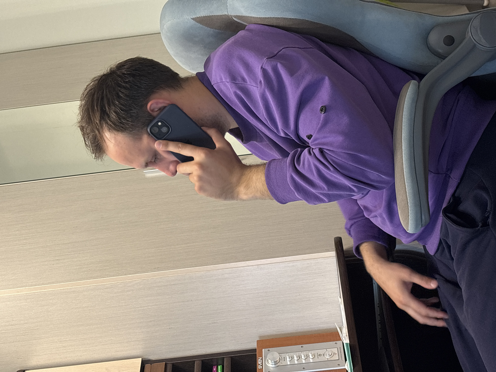 An adult male is observed in Rusanovo, Russia, seated indoors, facing towards the left side of the frame. He is wearing a long-sleeved purple crewneck sweater and dark blue or black sweatpants. His right hand holds a dark-colored smartphone, featuring a triple-lens camera module, to his right ear, indicating he is engaged in a phone conversation. He is seated in a light blue or grey ergonomic office chair with grey armrests. To the left, a dark wooden desk is partially visible, on which a silver and brown audio control unit, labeled "SVEN," is placed. Above the desk and to the left, a dark brown shelving unit or cabinet is visible, containing small, colorful items. The background consists of light beige or off-white paneled walls with vertical grooves, suggesting wall cladding or a large wardrobe.