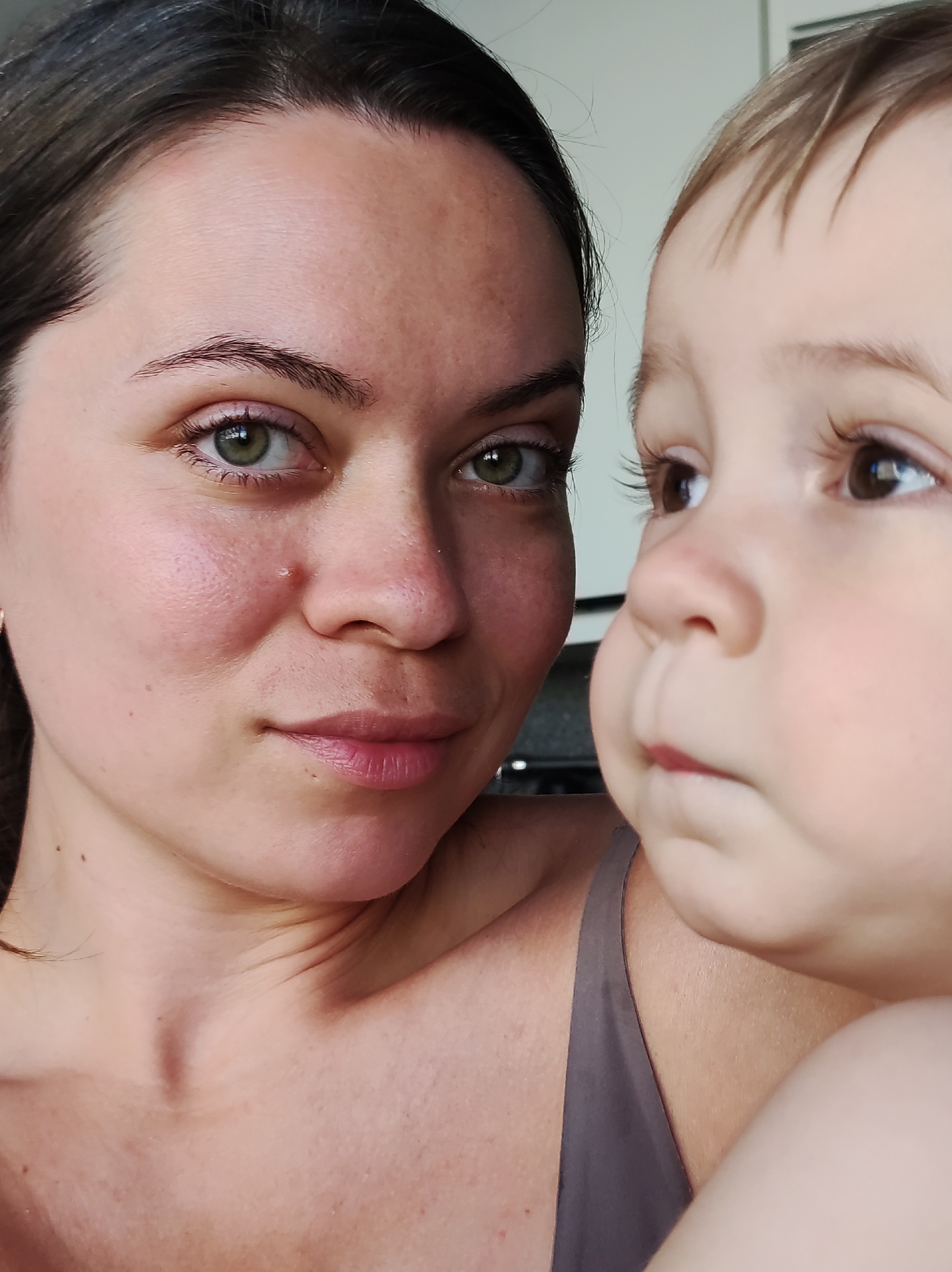 A close-up, medium-shot photograph features a woman and a young child. The woman, likely in her late 20s to mid-30s, faces the camera with a slight smile. Her hair is dark, pulled back from her face, and her eyes are green. The child, seemingly a toddler, is positioned slightly to the right of the woman and looks upwards and to the right, away from the camera. The child has light brown hair and dark brown eyes. Both the woman and child have fair skin tones.
The woman is wearing a gray tank top. The background is slightly blurred and indistinct but appears to be an indoor setting, potentially a kitchen or living space.
There is no indication of the location in the city of Chaikovsky, Russia.