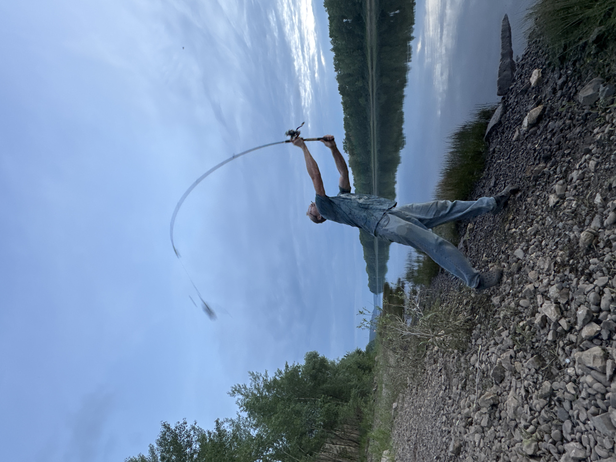 A person is fishing by a body of water. The person is standing on a rocky shore, holding a fishing rod that is bent significantly, indicating a fish is on the line. The person is wearing a patterned shirt and blue jeans. The scene is a natural setting with trees lining the opposite bank of the water. The sky is cloudy, and the water reflects the sky and trees. The image appears to be taken during the late afternoon or early evening due to the soft light. There is no visible text in the image.