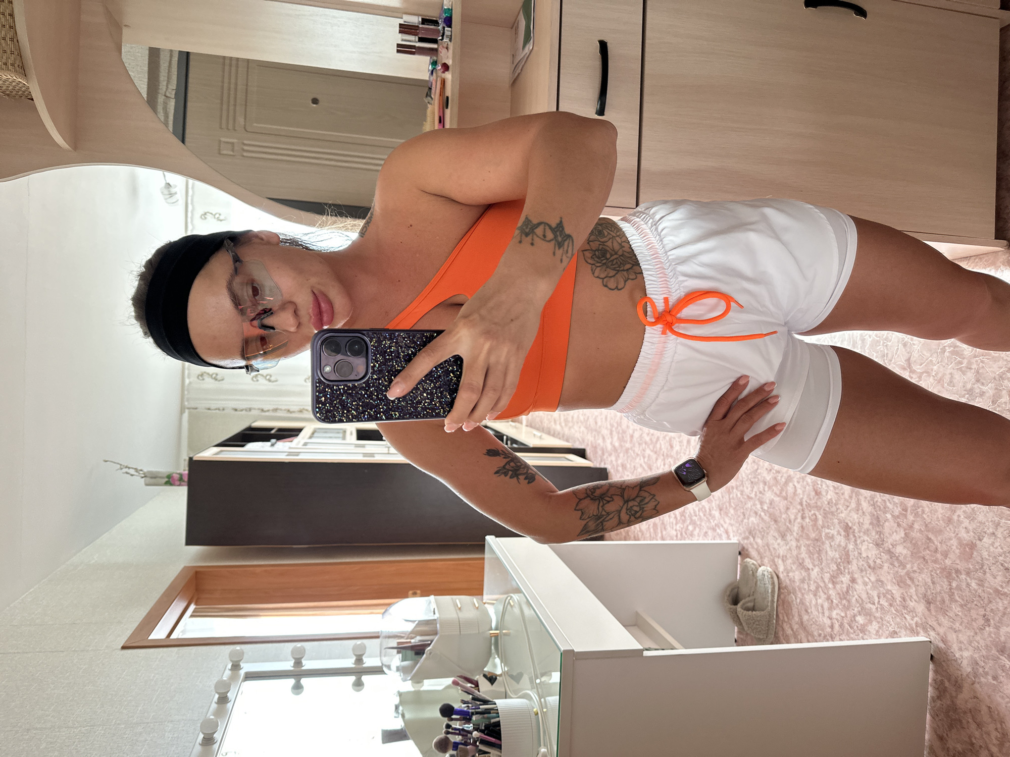 In a room with pink, textured flooring, a woman with a black headband and clear-lensed athletic glasses is taking a mirror selfie. She is wearing an orange sports bra and white athletic shorts with an orange drawstring. Tattoos are visible on her left arm and upper torso. On her left wrist, she wears a white smartwatch. To her left, a white vanity with a mirror adorned with lights and various makeup brushes and containers is partially visible. To her right, a closet with light wooden doors is present. In the background, a dark cabinet and part of a doorway are visible. The scene appears to be a personal dressing area or bedroom. The specific location in Yuzhnouralsk, Russia cannot be determined from the provided image.