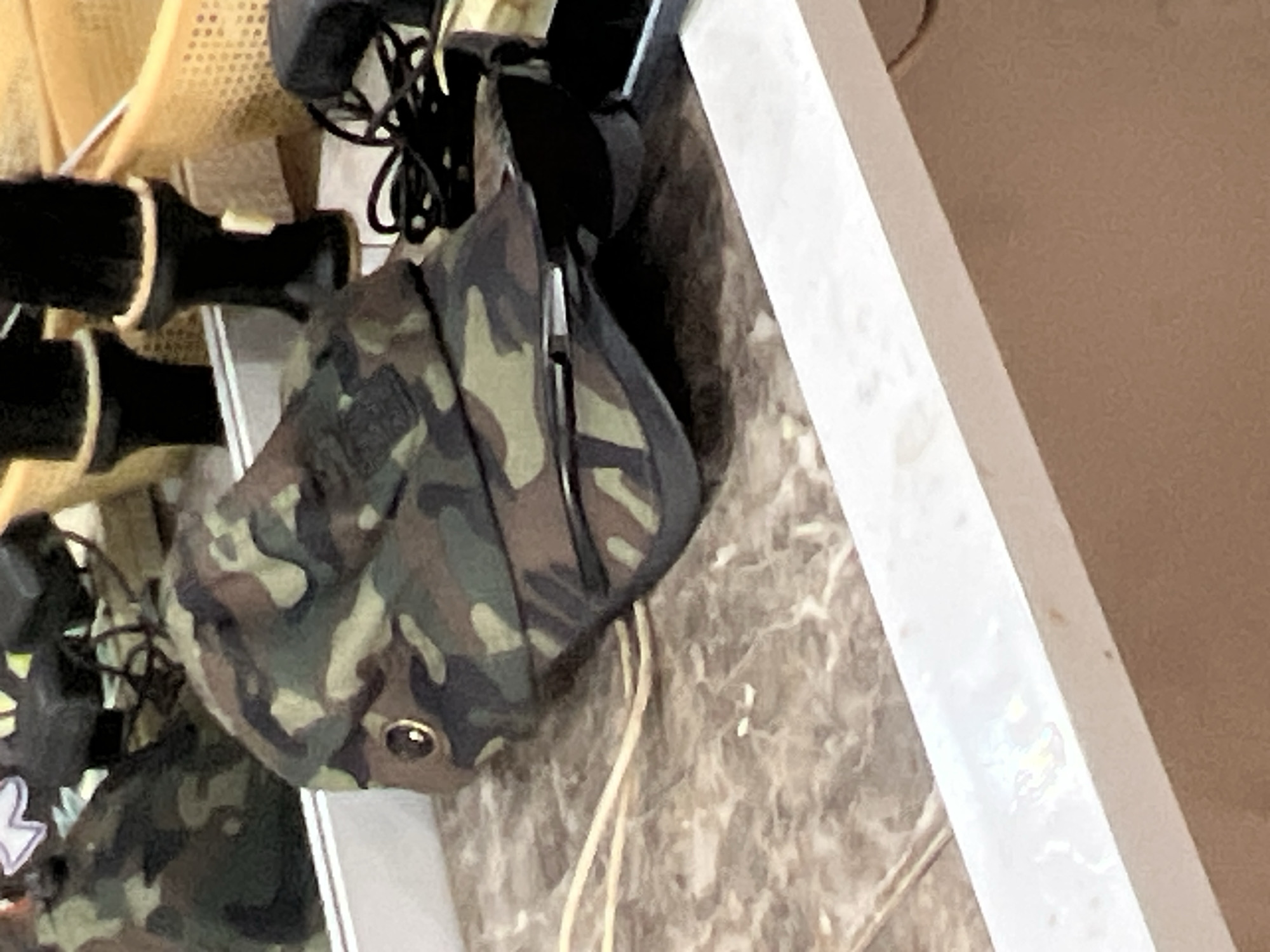 A camouflage-patterned baseball cap lies on a marble-patterned surface. The cap is tilted, with its brim pointing towards the right and its crown slightly elevated. A small brass-colored grommet is visible on the left side of the cap. In the background, there are reflections in a mirror of what appear to be black cylindrical objects with cream-colored bands, and a woven yellow basket. A black electronic device with a coiled cord is positioned behind the cap. The scene is indoors, likely in a domestic or retail setting. There are no people visible in the image. The location in Jalingo, Nigeria, is not discernible from the visual information provided.