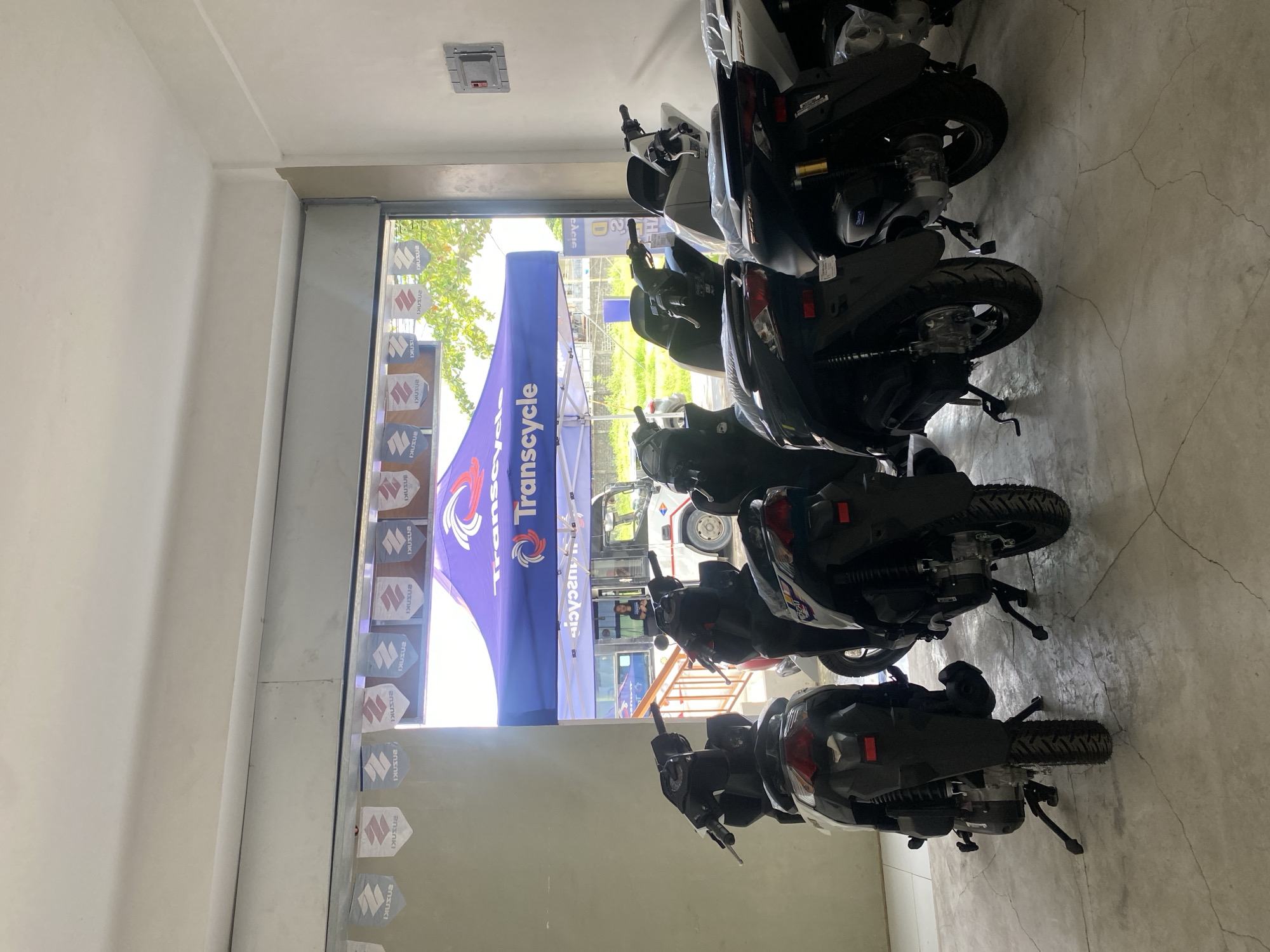 The image shows a Suzuki dealership in San Jose del Monte, Philippines. Several motorcycles are parked inside the showroom, with a Transcycle tent set up outside. A person is visible standing near a white van in the background. The dealership appears to be located on a street with trees and greenery visible beyond the entrance.