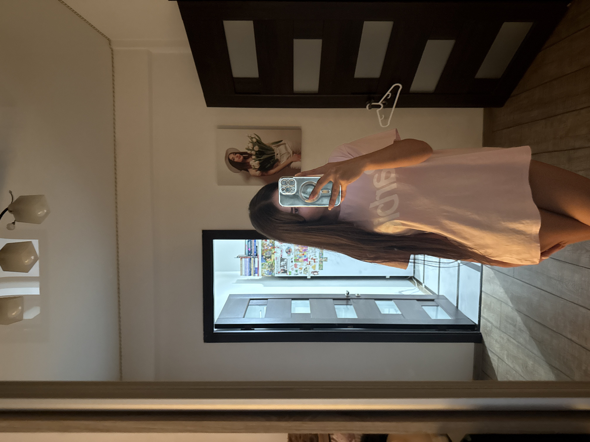 The image depicts a young woman taking a mirror selfie in a room. She is wearing a light pink t-shirt and has long dark hair. The room has wooden flooring and a large mirror on the left wall. To the right of the woman, there is a dark brown door with several horizontal panels and a white hanger on it. On the wall between the mirror and the door, there is a framed picture of a woman holding a bouquet of white tulips. Behind the woman, an open door leads to a hallway with a tiled floor and a refrigerator covered in various stickers. The overall lighting suggests it is daytime, with artificial light sources also visible on the ceiling. There are no specific landmarks or contextual clues within the image that would allow for its precise location within Chișinău, Moldova.