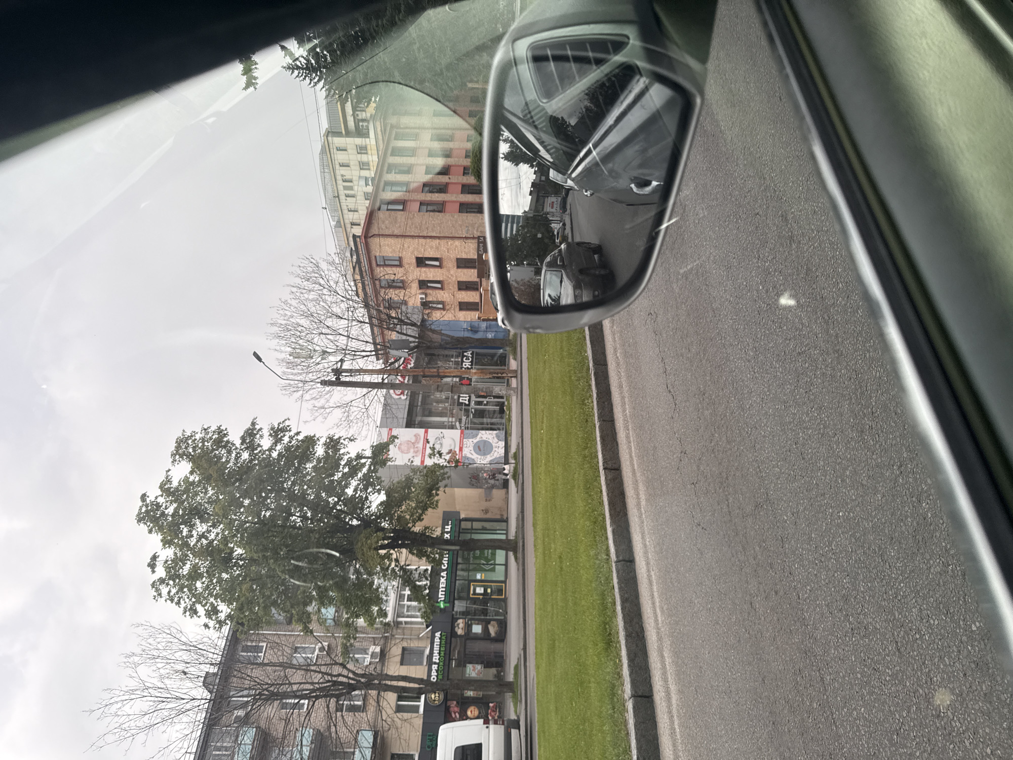 The image appears to have been taken from inside a vehicle, likely a car, while it is stopped on a street in Dnipro, Ukraine. The view out the window is towards the street and buildings. A side mirror of the car is visible in the upper right corner of the frame, reflecting the street behind the vehicle, including other cars. The street has a grassy median. Buildings of varying heights and architectural styles line the street, including commercial buildings with signs and storefronts. One visible storefront seems to be a pharmacy ("АПТЕКА"). There are also apartment buildings with brick facades. Trees are present along the street, some with leaves and others bare. The sky is overcast, suggesting a cloudy day. A white van is also partially visible. The scene does not depict any discernible people or interactions, focusing instead on the static environment.