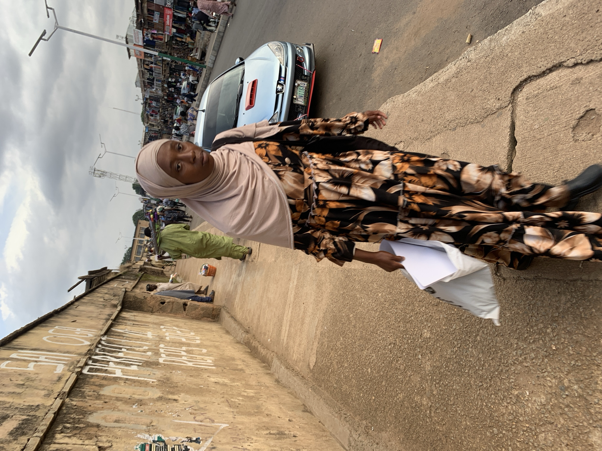 A woman in a light pink hijab and a brown and black floral patterned dress walks down a paved path. She is carrying a white plastic bag with papers inside. In the background, a light blue car is parked, and a street market with many stalls and people is visible. The sky is overcast, suggesting a cloudy day. The woman appears to be looking towards the camera. The scene is likely a street in Jos, Nigeria, given the context. The wall to the left has white graffiti on it.