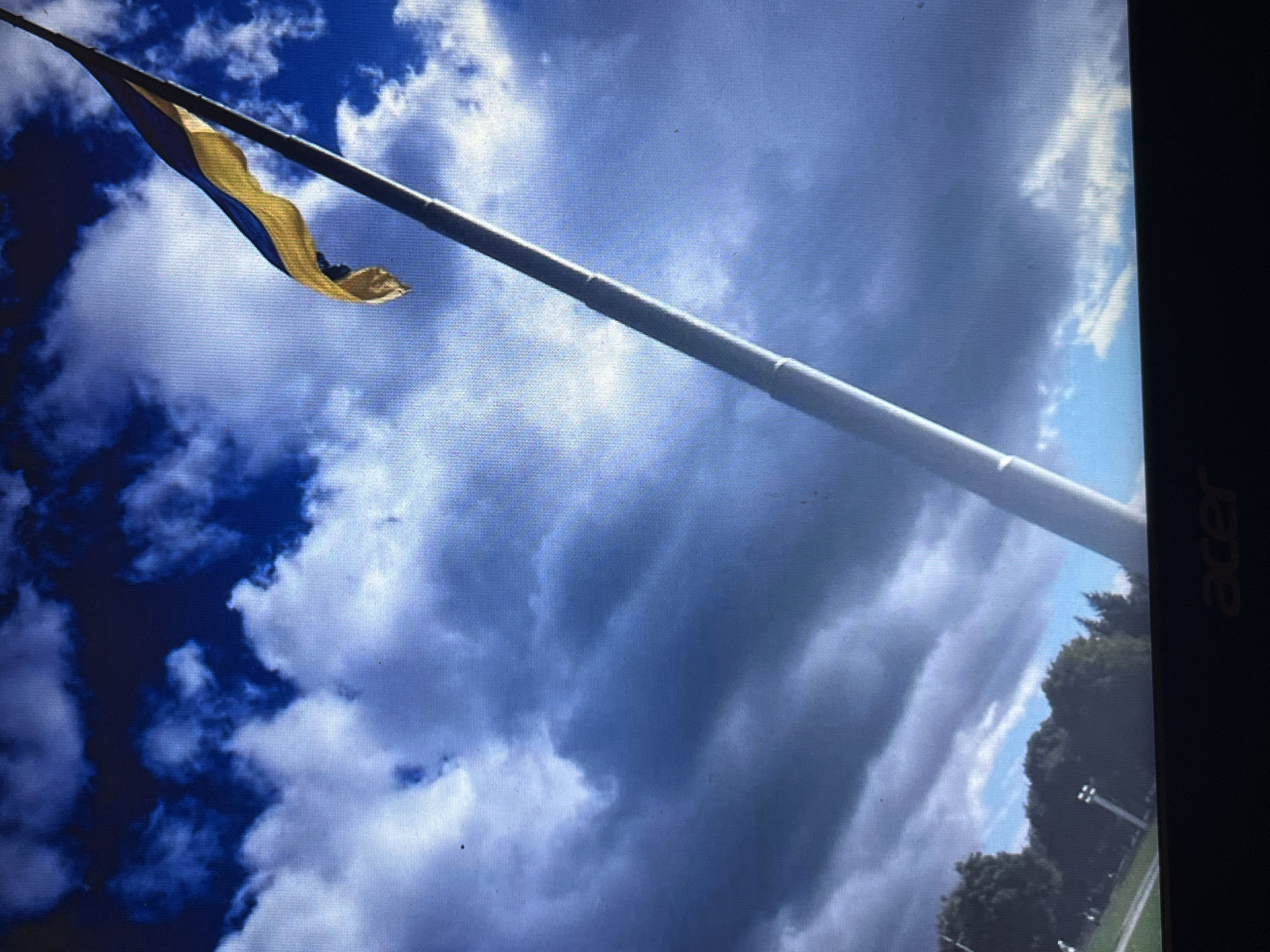 The image depicts a Ukrainian flag waving on a flagpole against a cloudy blue sky. The flagpole is tall and silver, extending diagonally across the frame. The flag, with its distinctive blue and yellow horizontal bands, is partially unfurled and caught by the wind. In the lower left portion of the image, trees and some lights can be seen, suggesting an outdoor setting in a park or open area. The bottom edge of the image shows the black bezel of a computer monitor with "acer" branding visible. The scene is an outdoor shot, likely taken during the daytime, with a focus on the flag and sky. The location of Bilenke, Ukraine, is not visually identifiable within the image itself.
