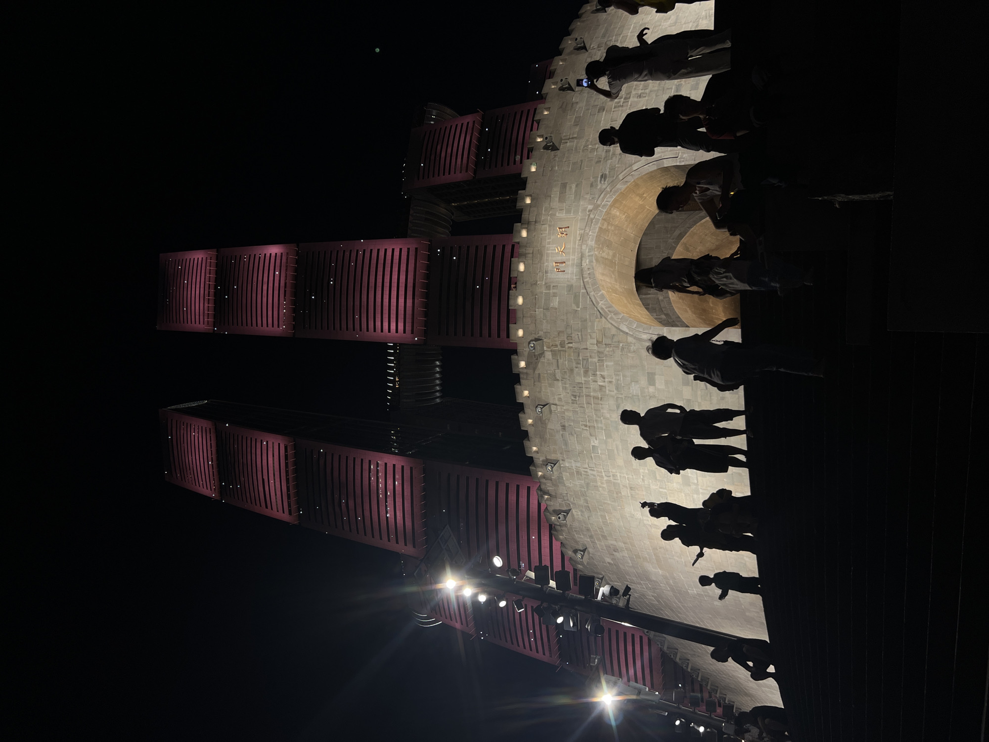 The image captures a night scene in the Yuzhong District of China, featuring a modern architectural marvel with a historical facade. Two towering skyscrapers, illuminated in a vibrant pink hue, dominate the upper portion of the frame. Their tiered structure and horizontal banding create a striking visual. Below the skyscrapers, a light-colored, fortress-like structure with crenellated battlements serves as a base. An arched gateway is visible in the center of this structure, with Chinese characters inscribed above it.
In the foreground, silhouetted figures of people are scattered across a wide set of dark stairs leading up towards the fortress. Some individuals are standing, while others are seated or crouching. Their poses suggest they are observing the illuminated buildings and the overall spectacle. One person is seen holding up a mobile phone, presumably to take a photograph. The lighting is dramatic, with the bright pink of the towers contrasting sharply with the dark night sky and the shadowed foreground. Streetlights and spotlights are also visible, casting a glow on the scene and creating lens flares. The overall impression is one of urban dynamism, where contemporary architecture is presented with a nod to historical aesthetics, attracting a crowd of onlookers.