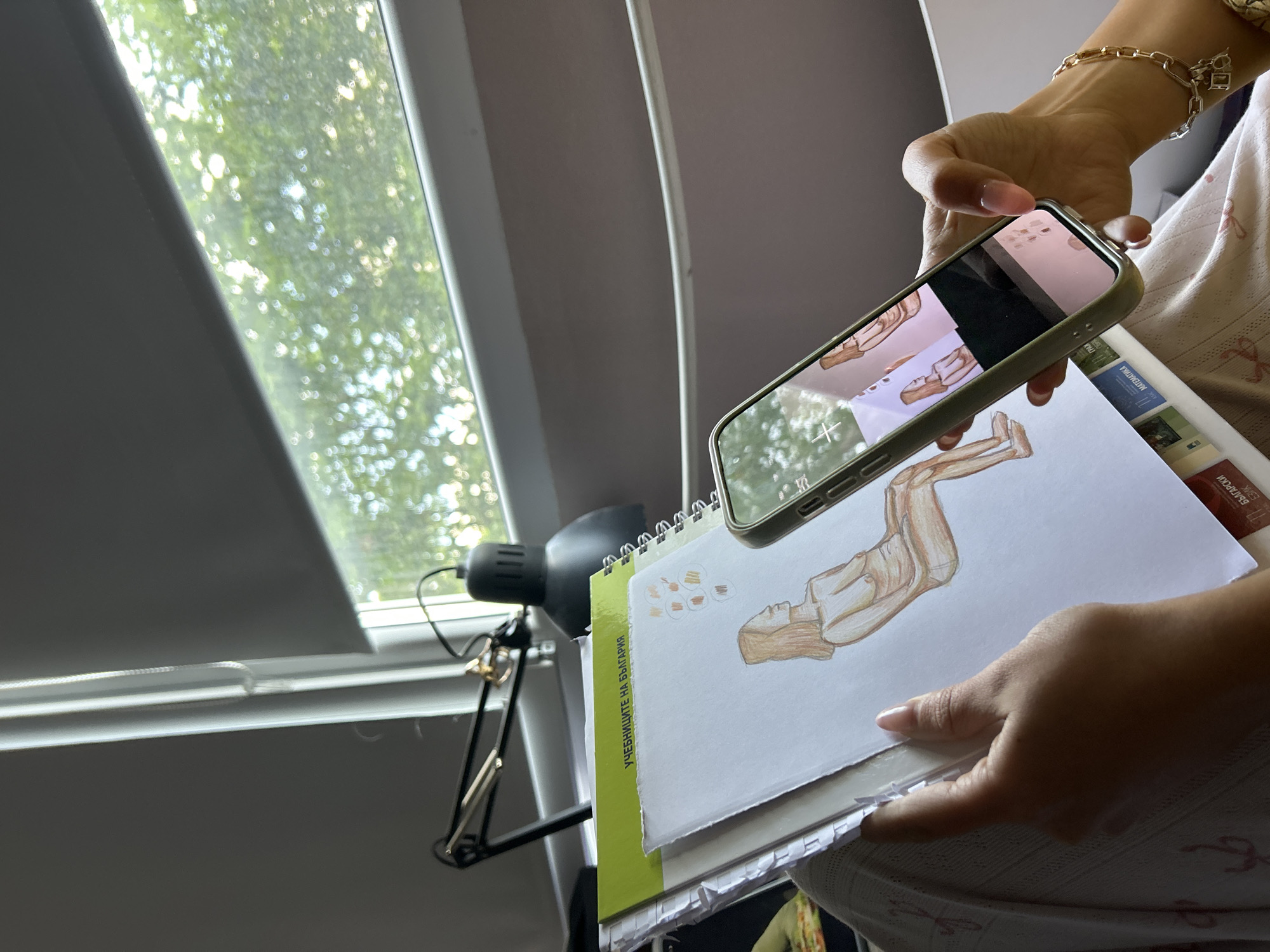 A person holds a spiral-bound notebook with a drawing of a figure, taking a photo of it with a smartphone. The phone's screen reflects the drawing and notebook pages. The notebook has a lime green spine labeled "УЧЕБНИЦИТЕ НА БЪЛГАРИЯ" (The textbooks of Bulgaria). Additional books are stacked underneath. The person wears a gold-tone bracelet and a printed garment. A black desk lamp is visible to the left of the notebook. A window with blinds is in the background, showing a view of green foliage.
There is no definitive evidence to suggest that the location is in the city of Sofia, Bulgaria.
